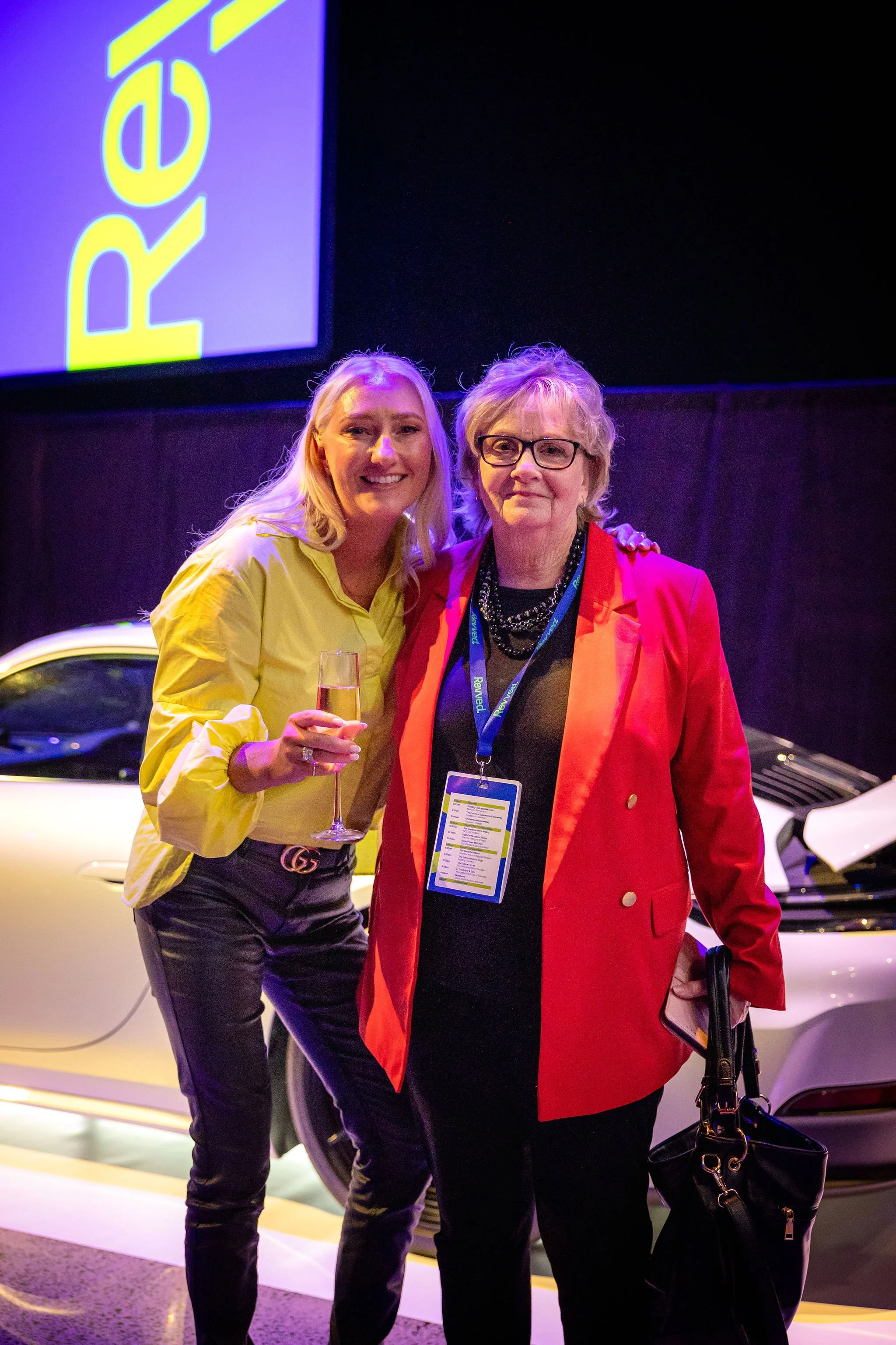 Two women standing next to a white car, smiling and posing for a photo at a car event or show. The woman on the left is holding a glass of champagne, wearing a yellow blouse and black pants. The woman on the right is wearing glasses, a red blazer, bl