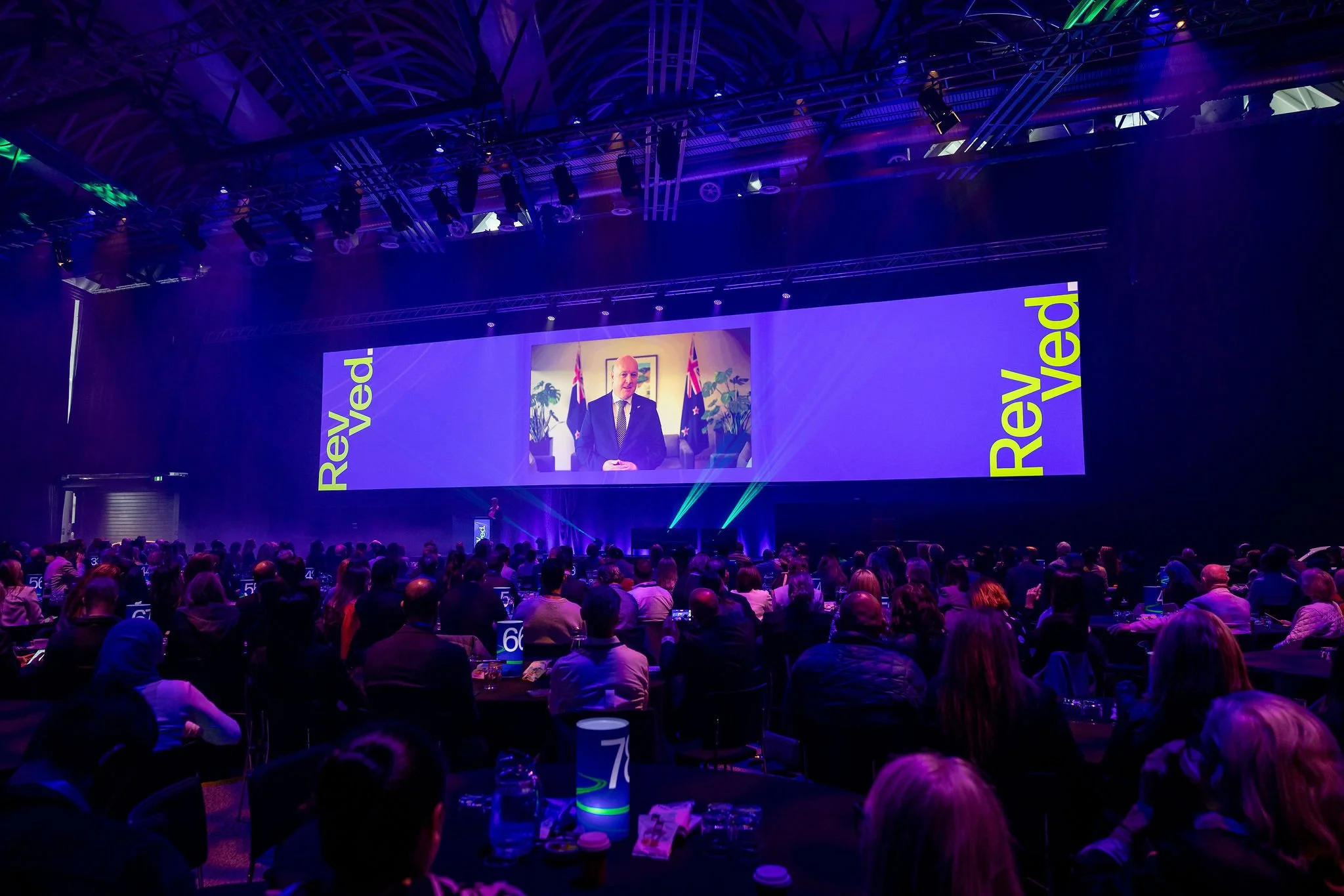 The Revved conference hall with many attendees seated at tables, watching a presentation on a big screen displaying a man in a suit with flags in the background.