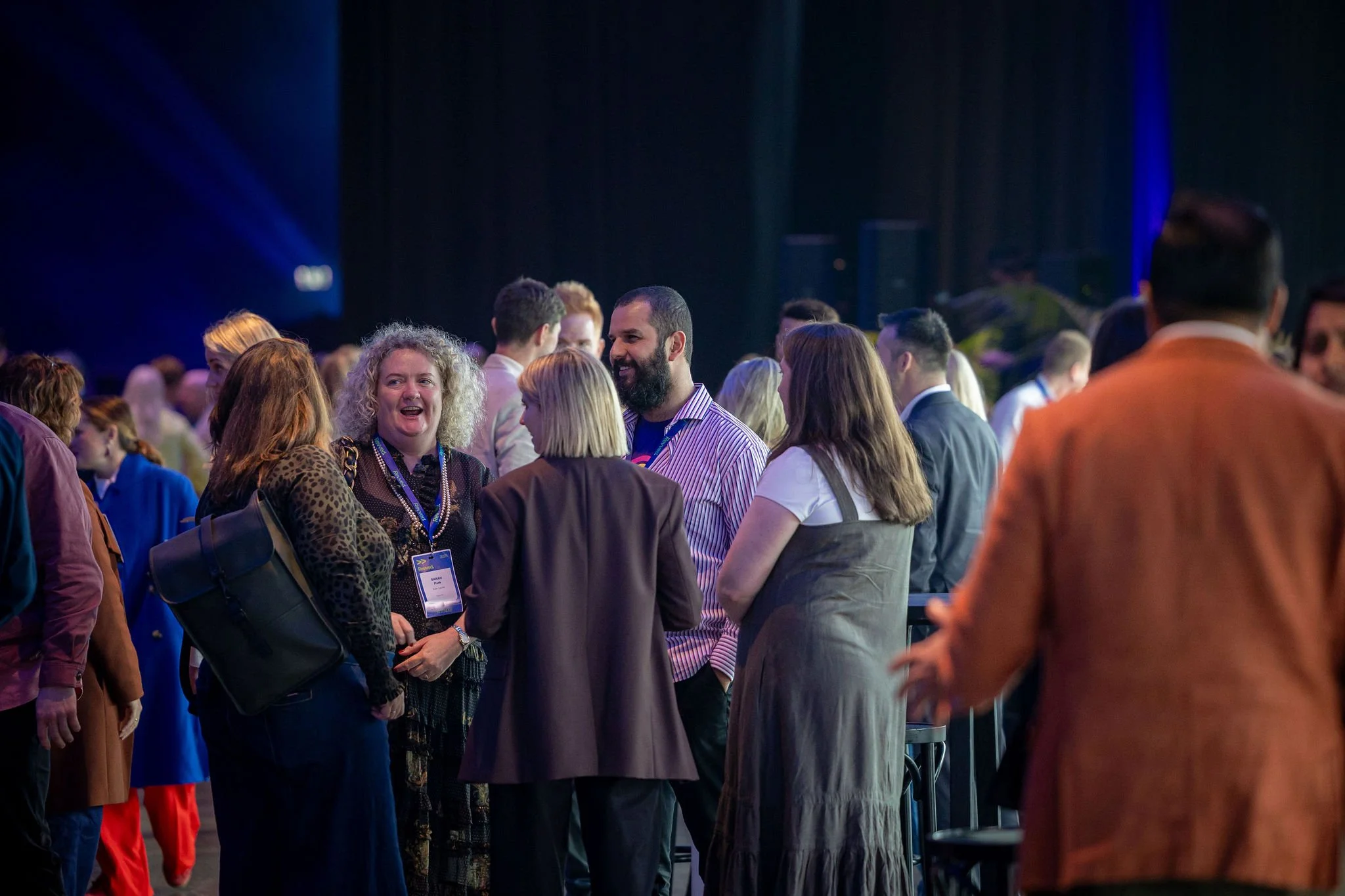People gathered at a conference, engaging in conversation, with some wearing conference badges, in a dimly lit room with blue and purple lighting.