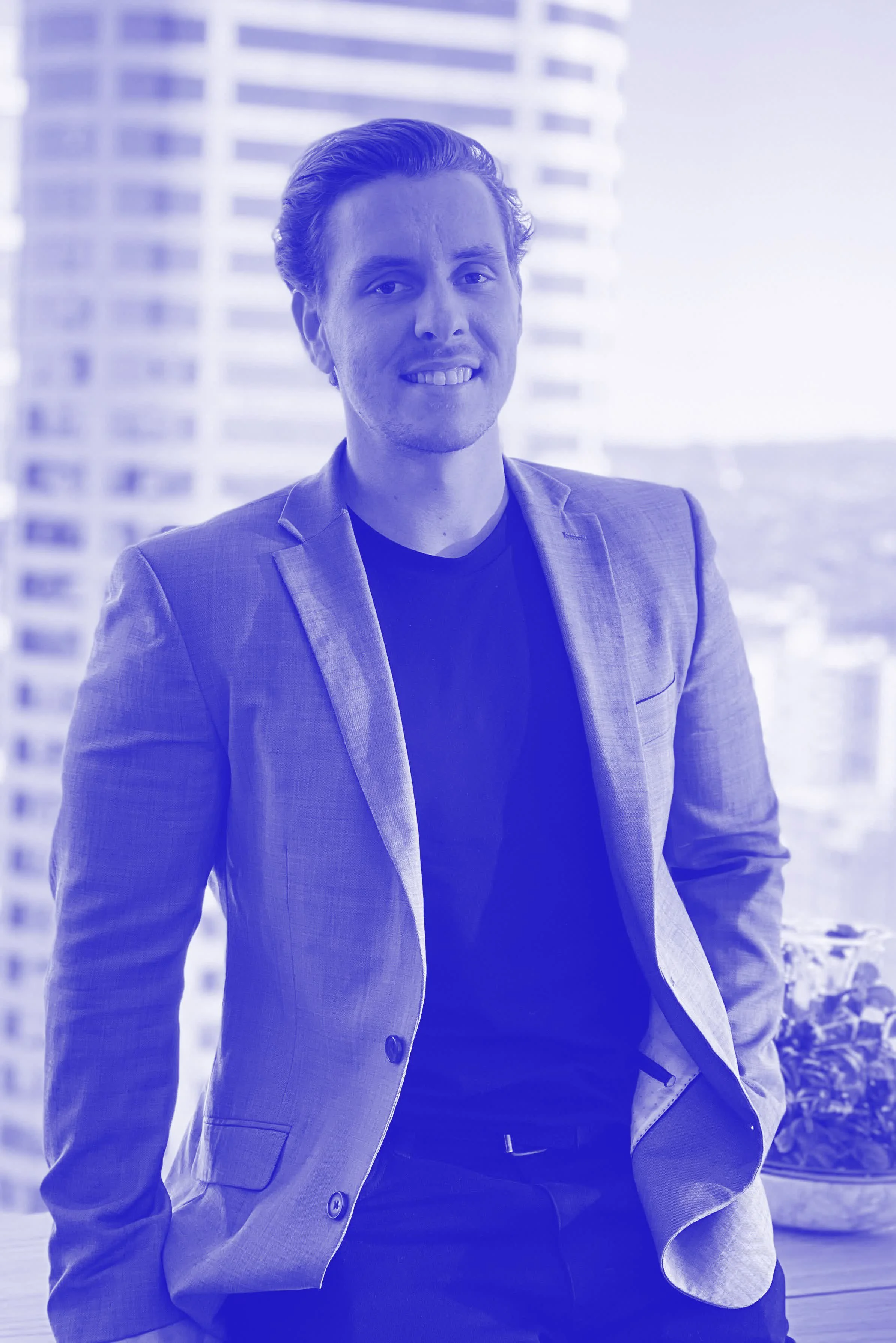 A man in a gray blazer and black shirt leaning against a railing with a city skyline in the background.