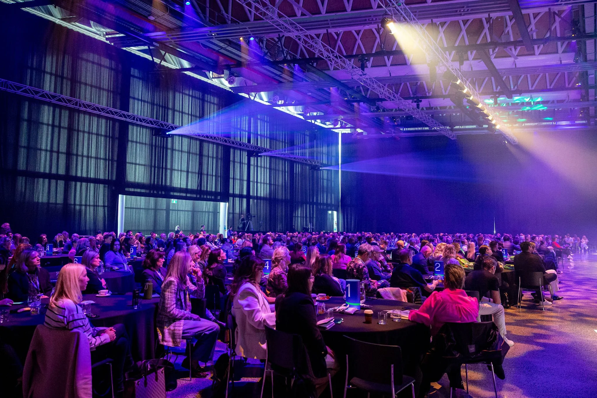 Revved attendees seated at tables in a large conference hall with purple and yellow stage lighting, high ceiling, and industrial beams.