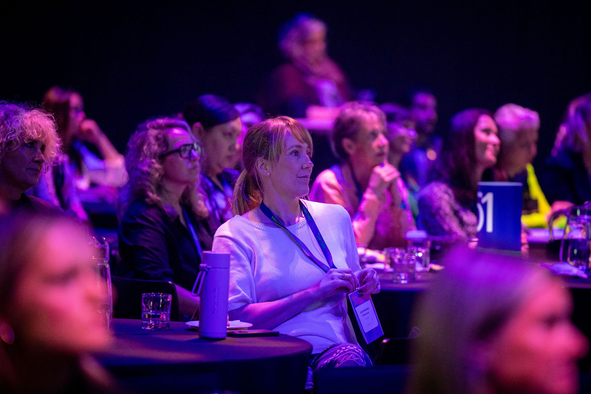 Audience of predominantly women at Revved, sitting at tables in a dark, purple-lit venue, focused on a speaker or presentation