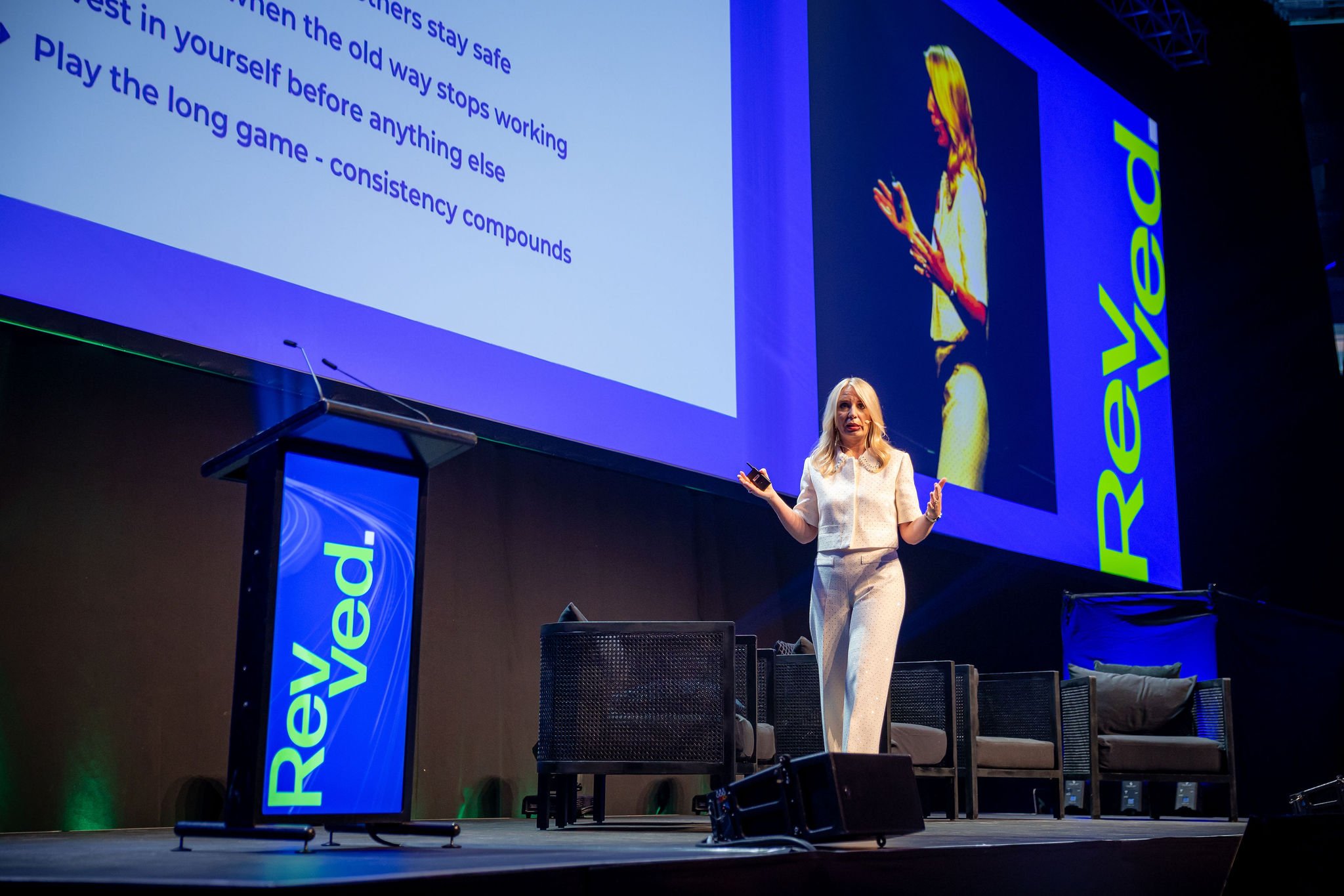 A woman with blonde hair giving a presentation on a stage at a conference. Large screens behind her display slides with text, including phrases like 'stay safe', 'old way stops working', and 'long game - consistency compounds'. The stage features bra
