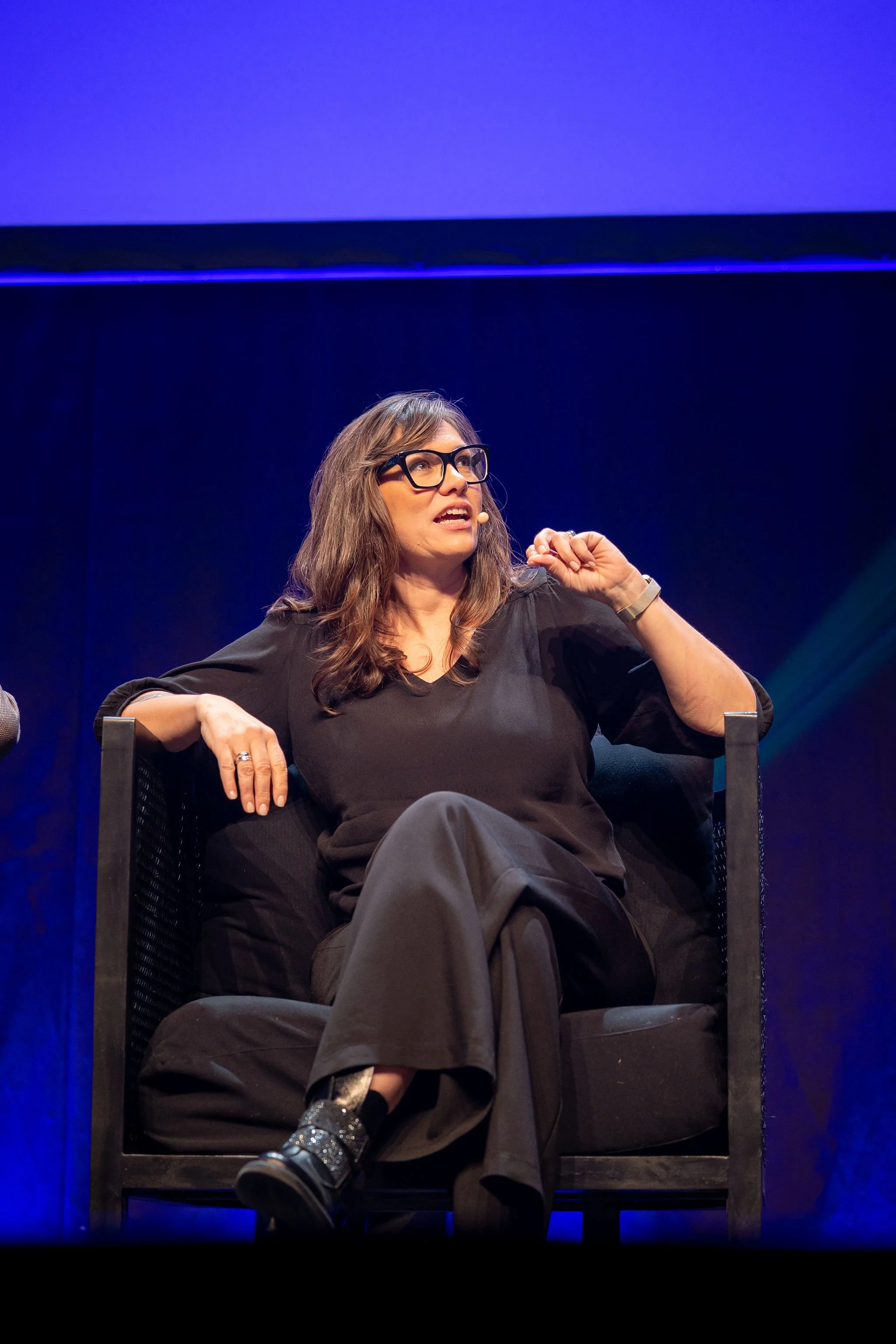 A woman sits presenting on stage at Revved with a dark background and purple lighting. 