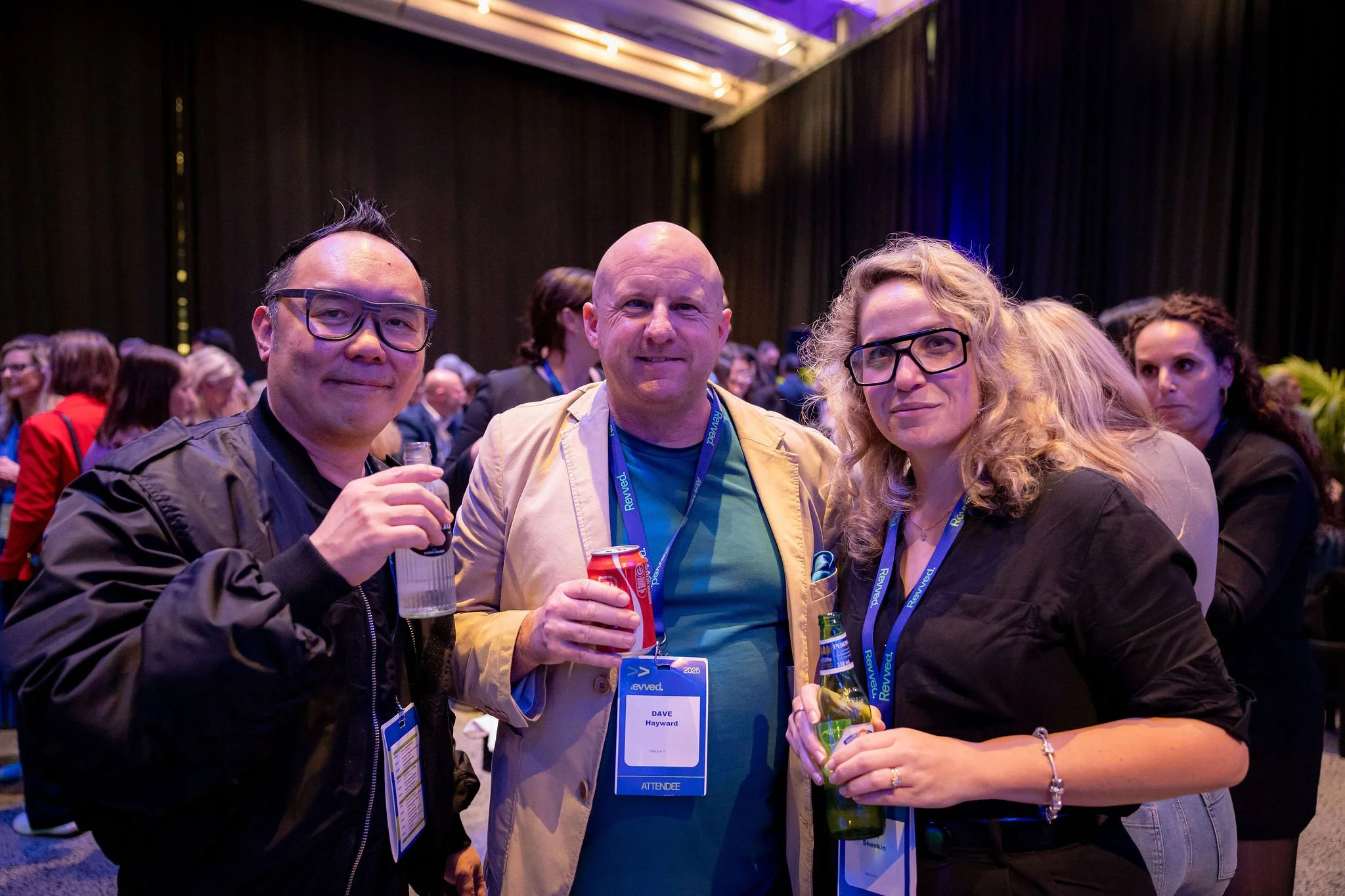 Three people, two men and a woman, standing close together at a conference or event, holding drinks, smiling, with a dark curtain background and other attendees visible behind them.
