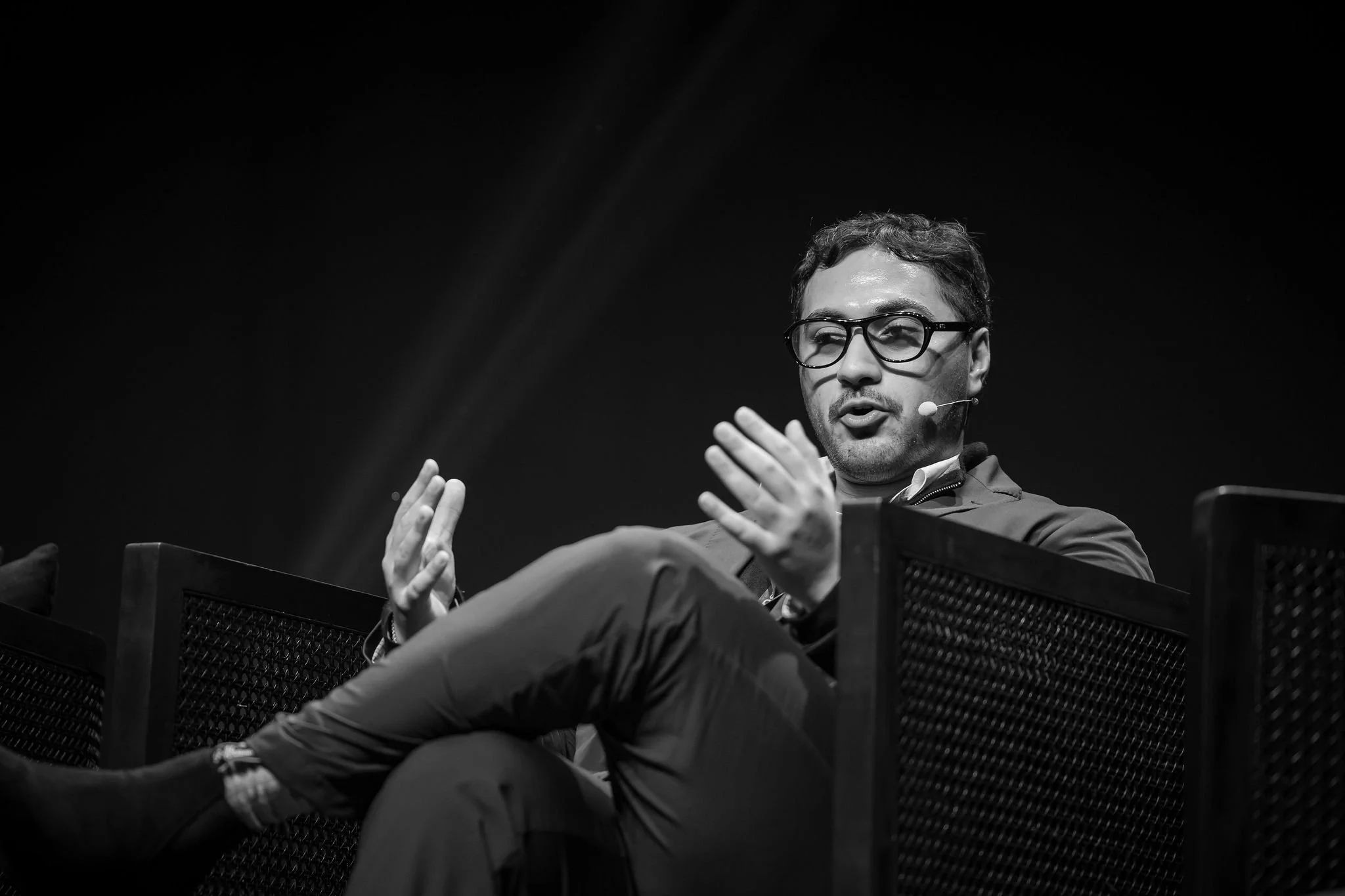 A man with glasses and a headset speaking, sitting on a stage with a dark background.