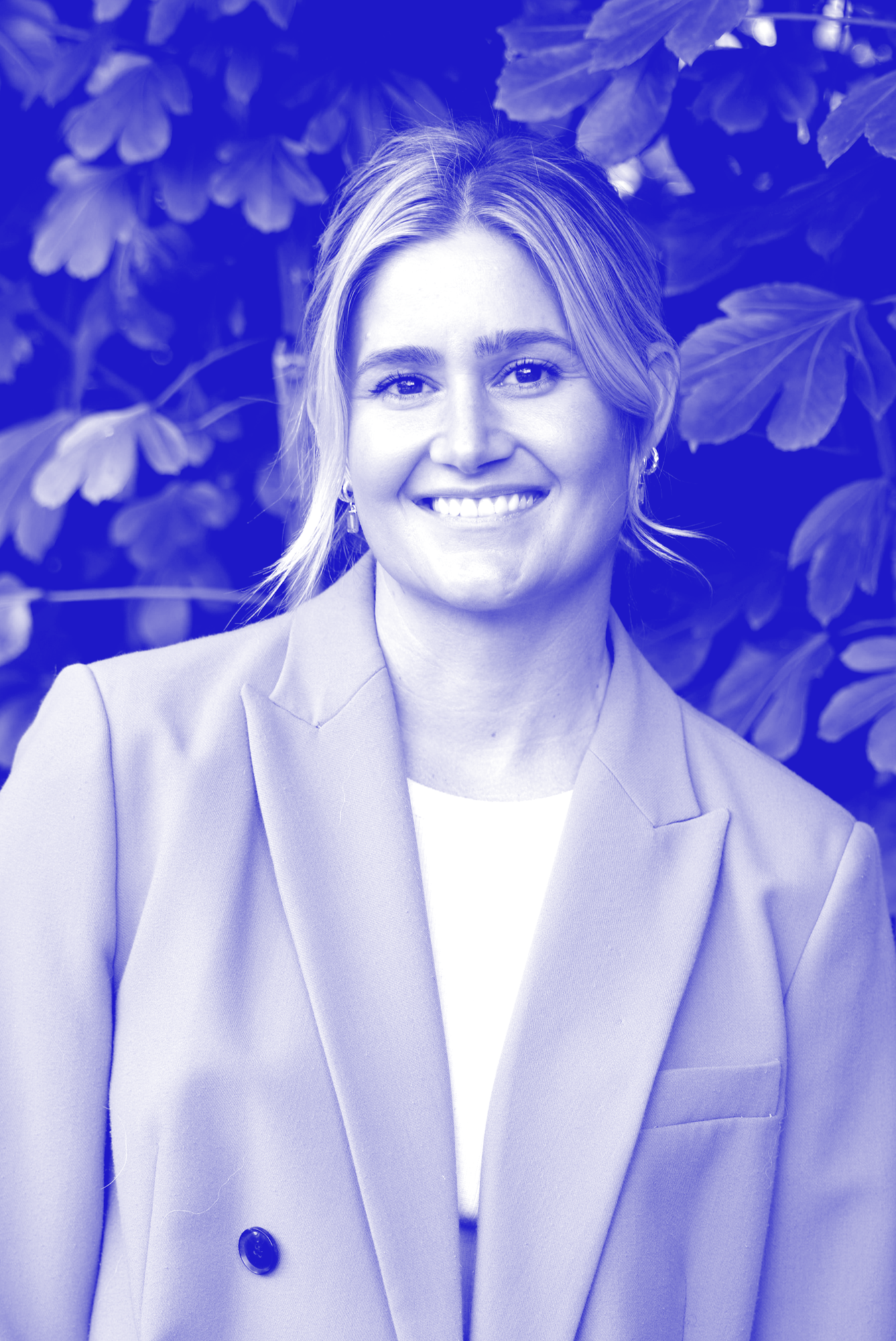 Close-up of a woman with blonde hair smiling, wearing a blazer and earrings, standing in front of leafy greenery.