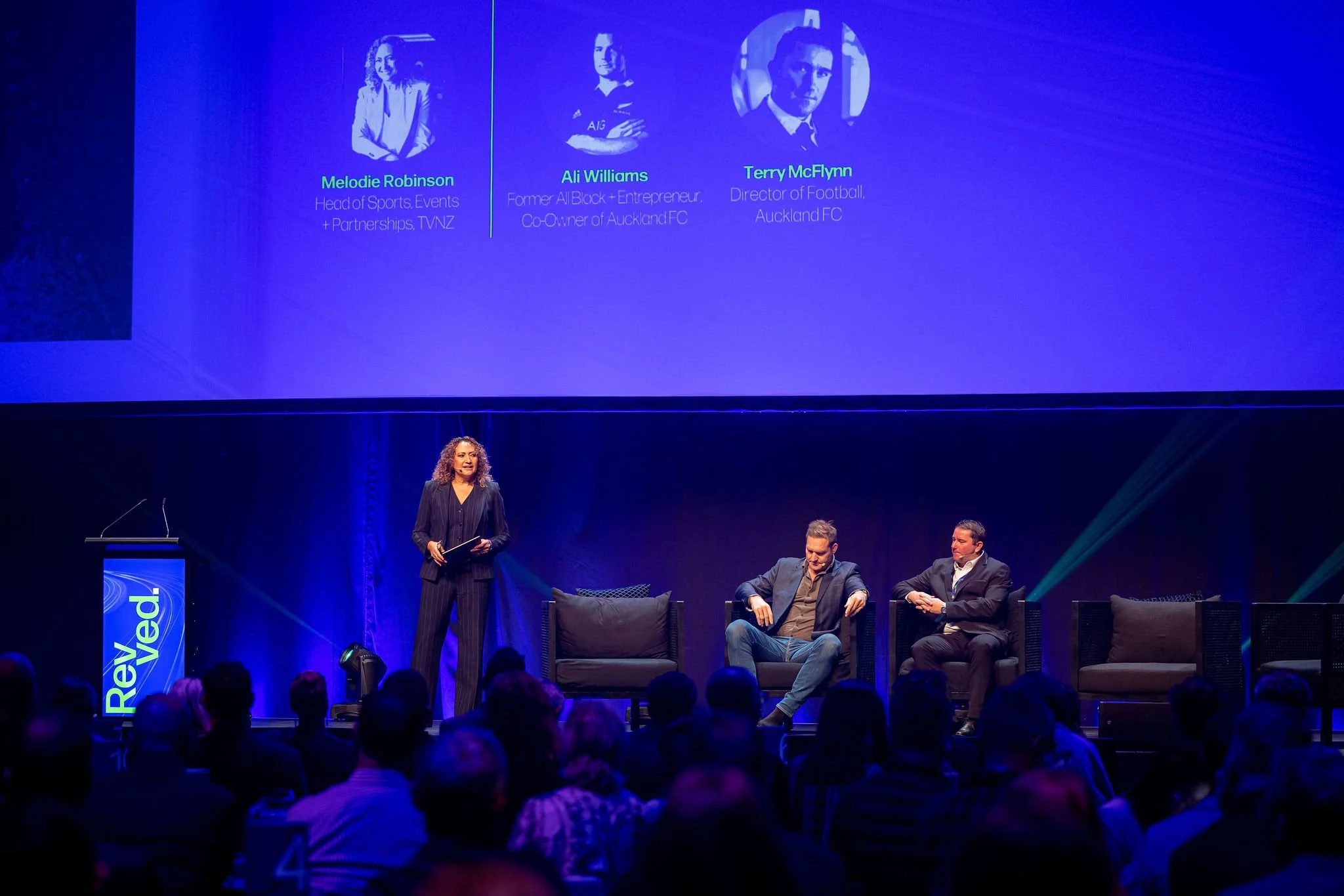 A woman on stage at Revved speaking into a microphone while three men sit on chairs behind her in front of an audience. A large screen displays three headshots with names and titles.