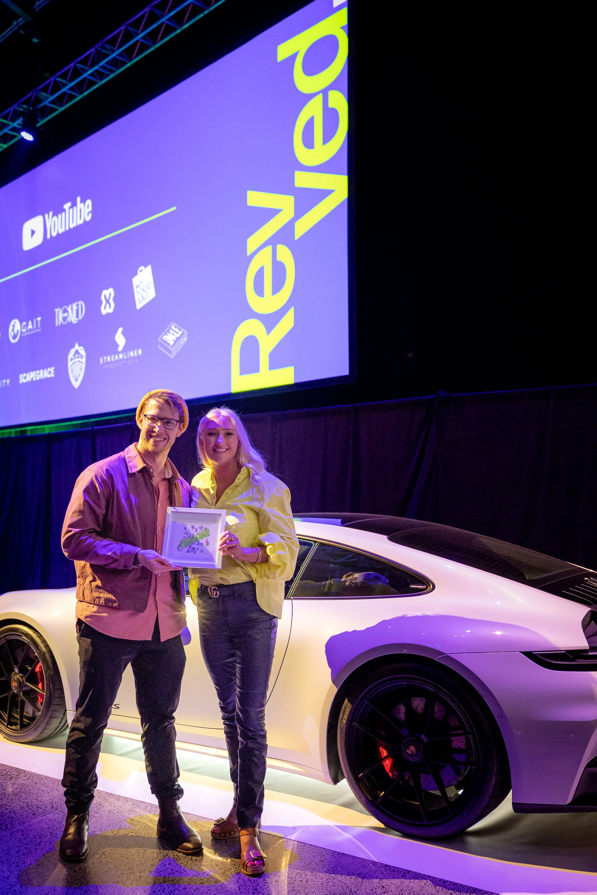 Two people smiling and holding a framed picture in front of a white luxury sports car at an event with a large screen displaying the YouTube logo and various icons in the background.