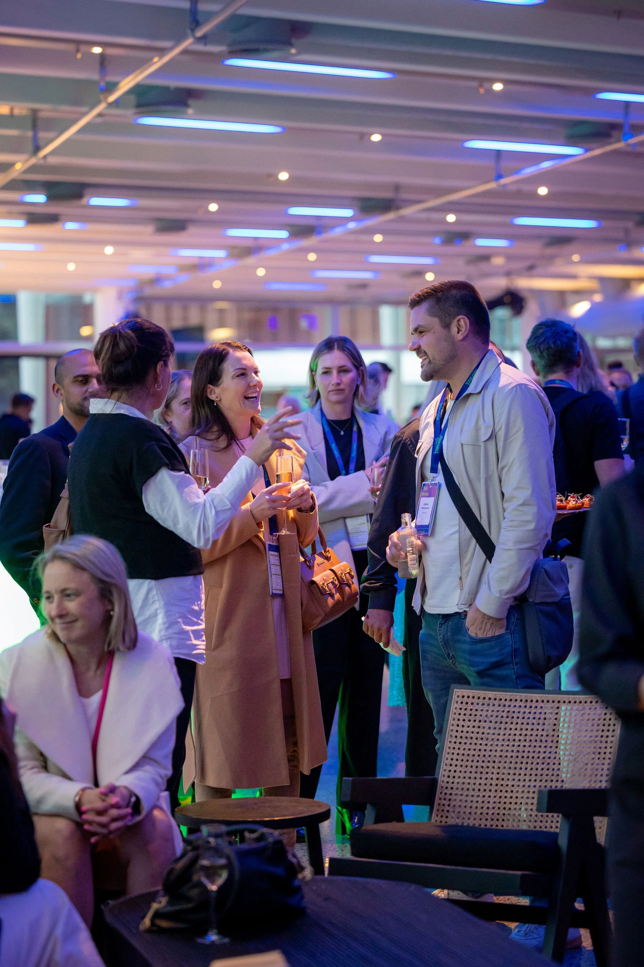 People socializing at a networking event with drinks, in a modern indoor venue with a ceiling illuminated by blue lights.