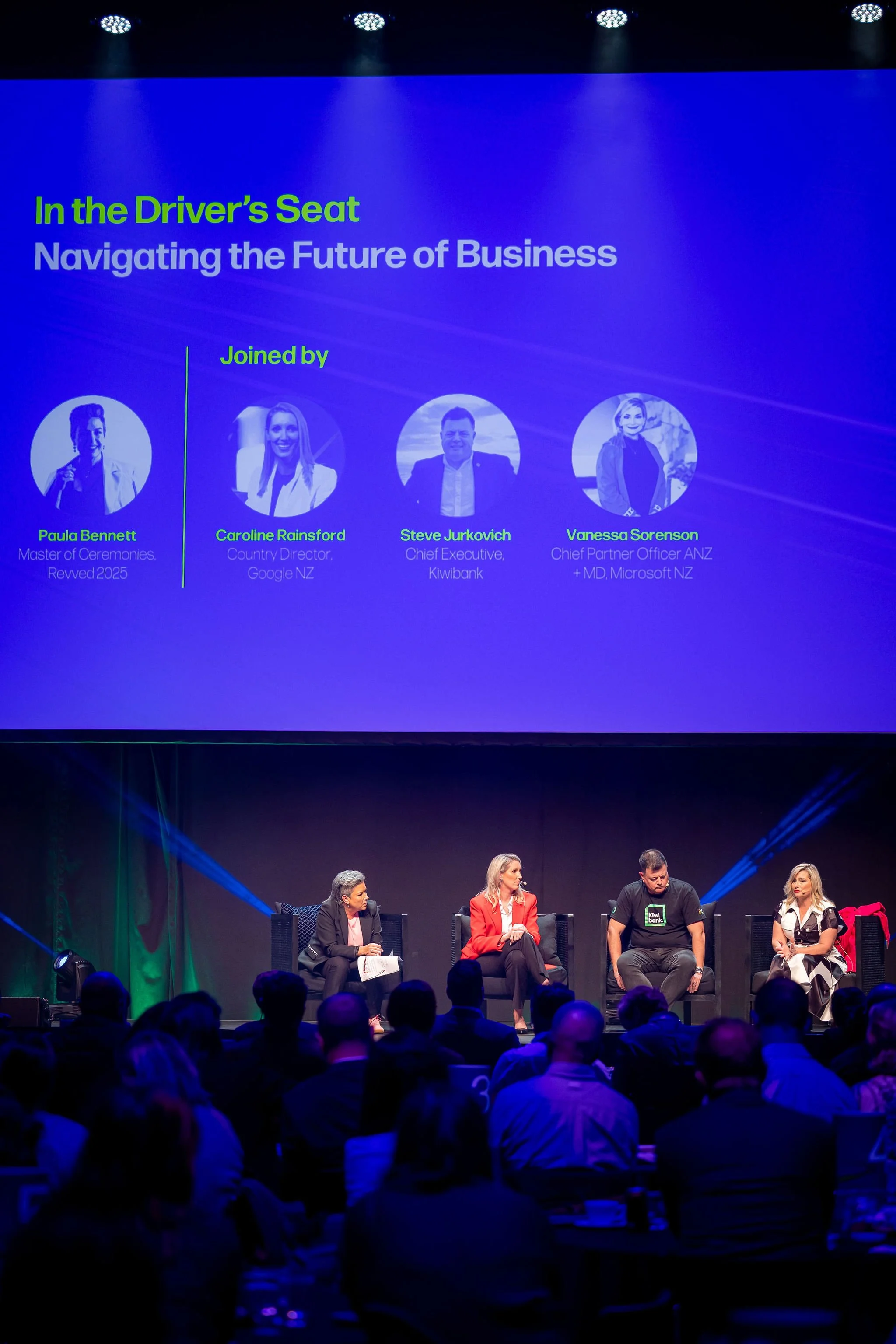 A panel discussion is taking place on stage with four women seated in chairs. Behind them is a large screen displaying a presentation slide with a blue background and images of the speakers, including their names and titles. The audience is visible i