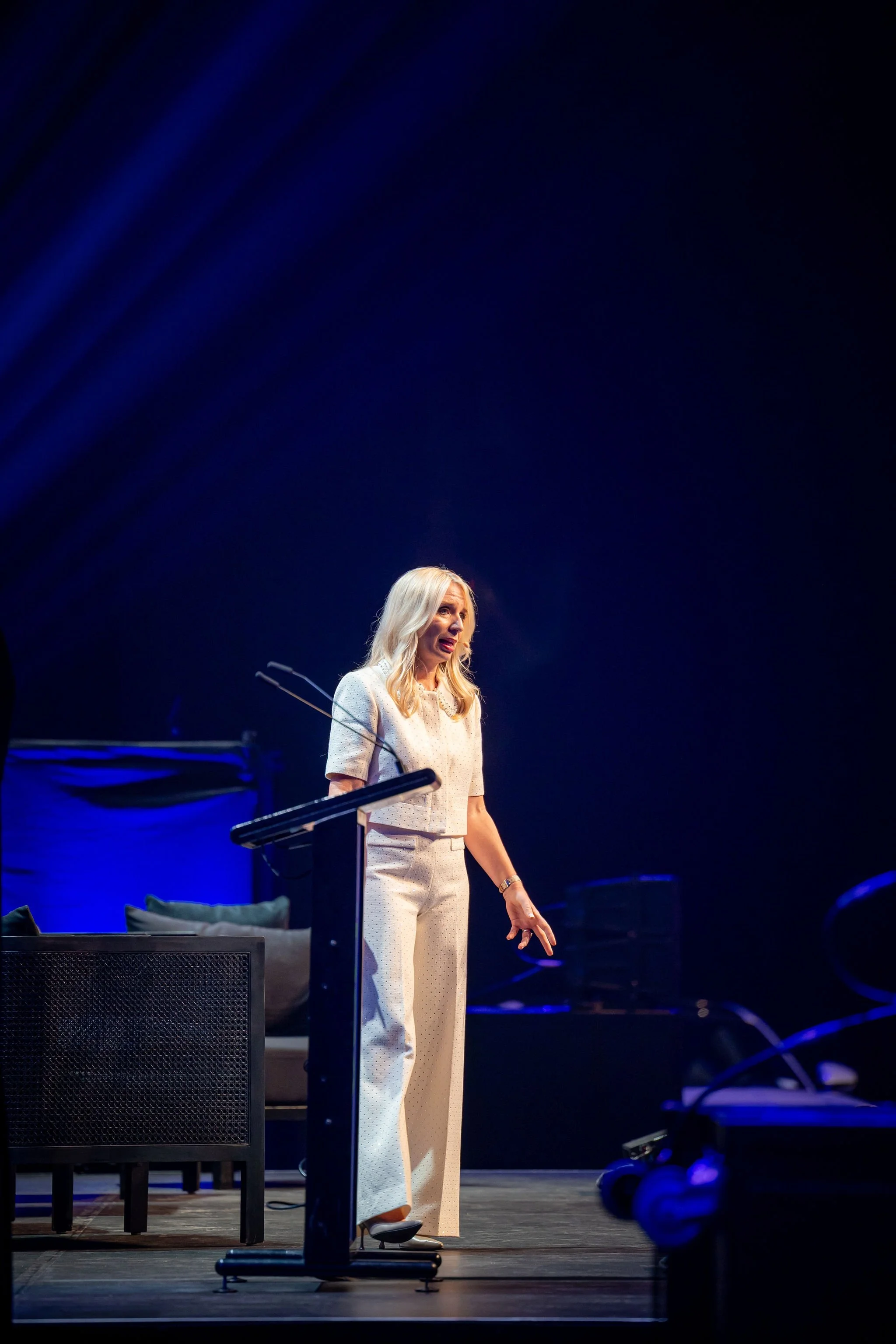 A woman with long blonde hair is speaking at a podium on a stage with a dark blue background. She is wearing a white outfit with a textured pattern and is gesturing with her right hand.