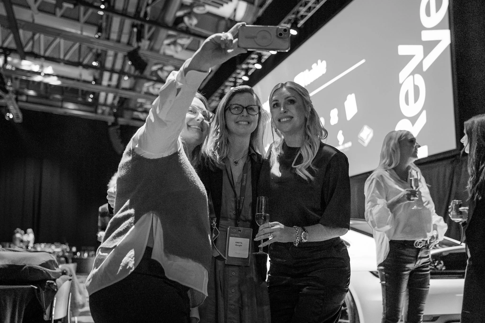 Three women at a social event taking a selfie together, all holding wine glasses, in a large indoor venue with a high ceiling and an LED screen in the background.