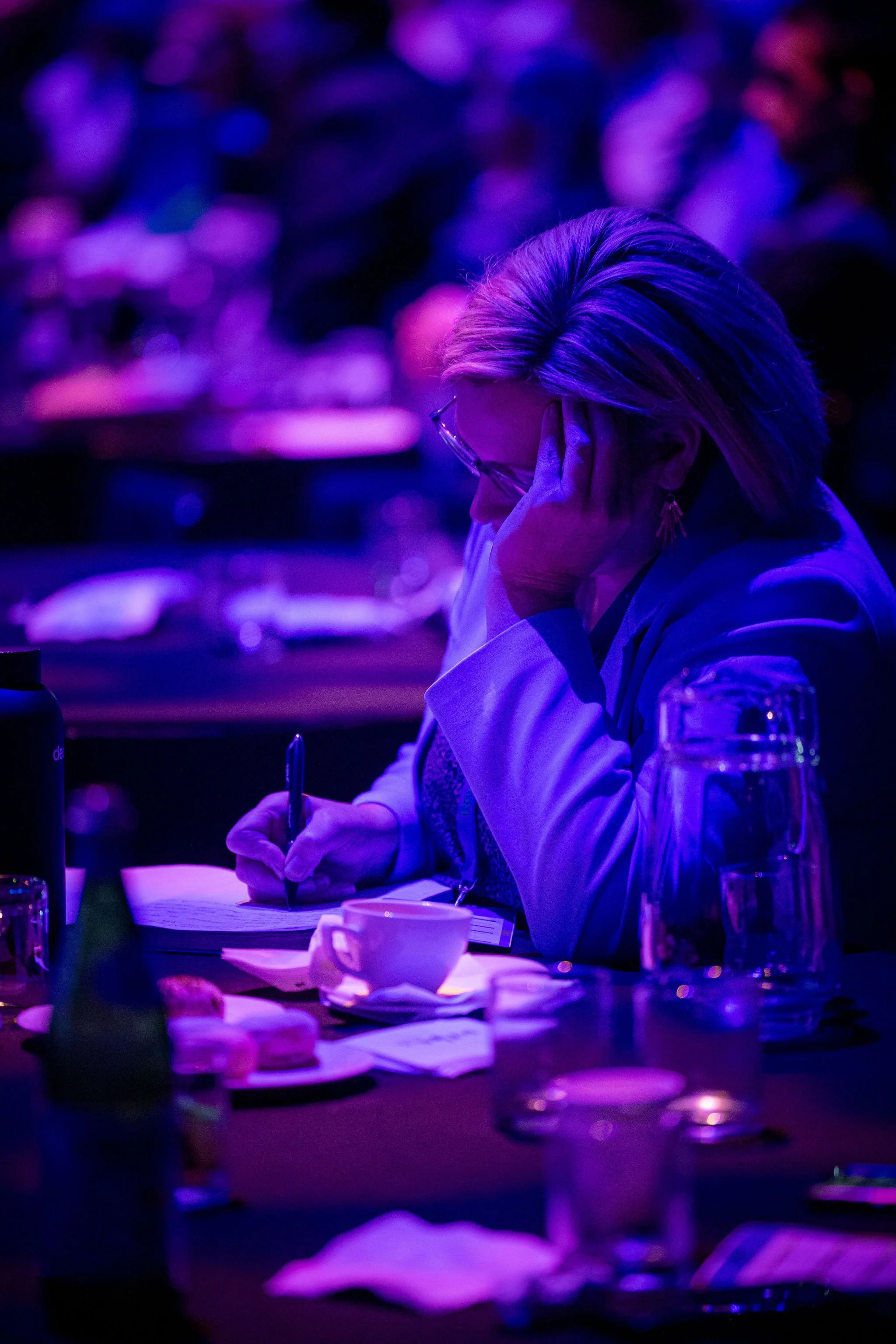 A woman with short blonde hair and glasses is sitting at a table, writing on paper with a pen, under purple lighting with glasses and a cup on the table.