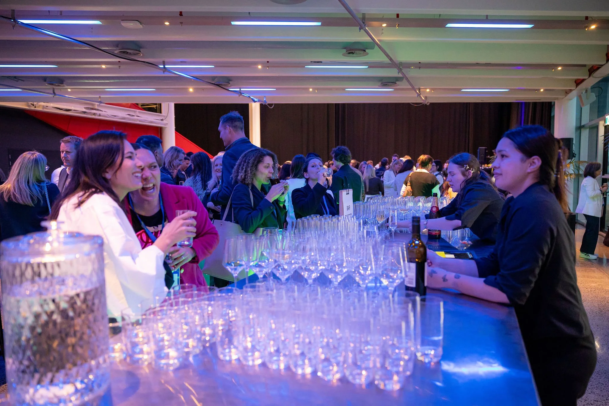 People socializing and drinking at a bar or event space with a long table and illuminated ceiling lights.