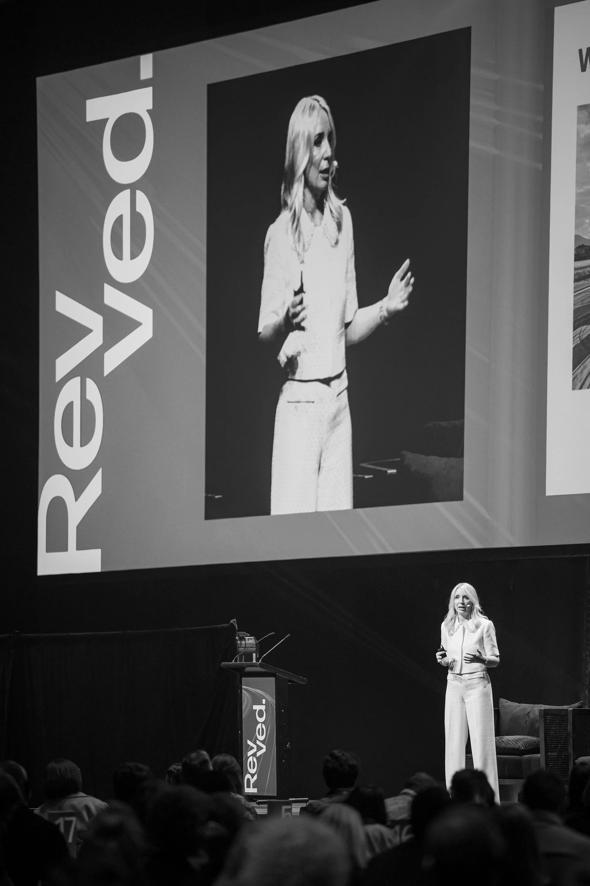 A woman is giving a presentation on stage with a large screen behind her showing her image. She is wearing a light-colored blouse and wide-leg pants, holding a remote control, and speaking to an audience.