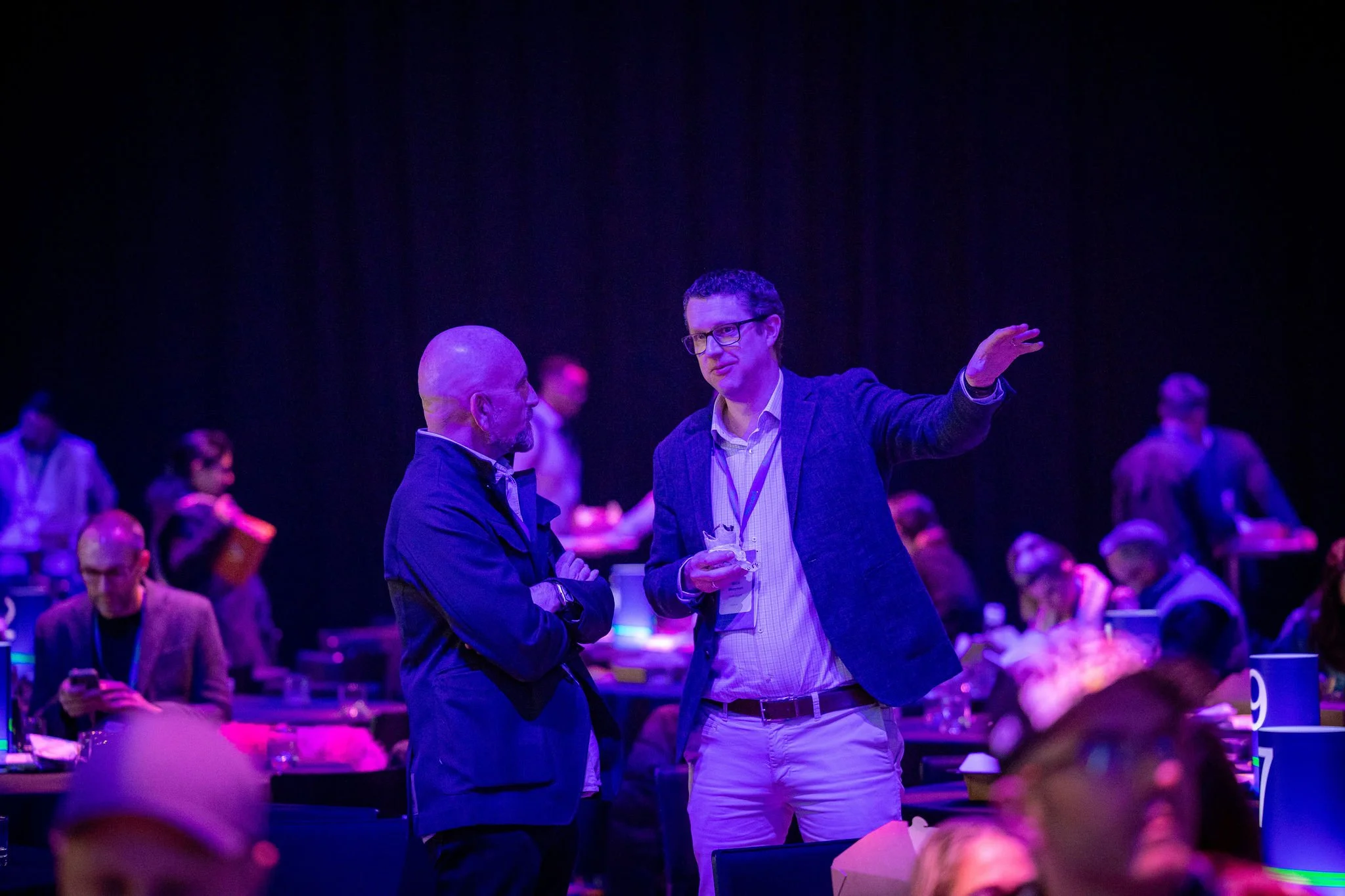 Two men are talking at a conference or seminar, illuminated by purple and blue lighting, with many other attendees seated around them.