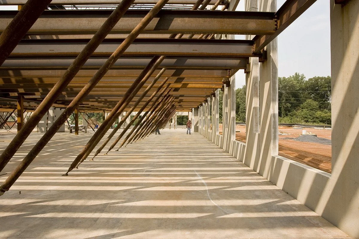 Construction site with a partially built structure, showing steel beams and concrete flooring, with workers in the background.