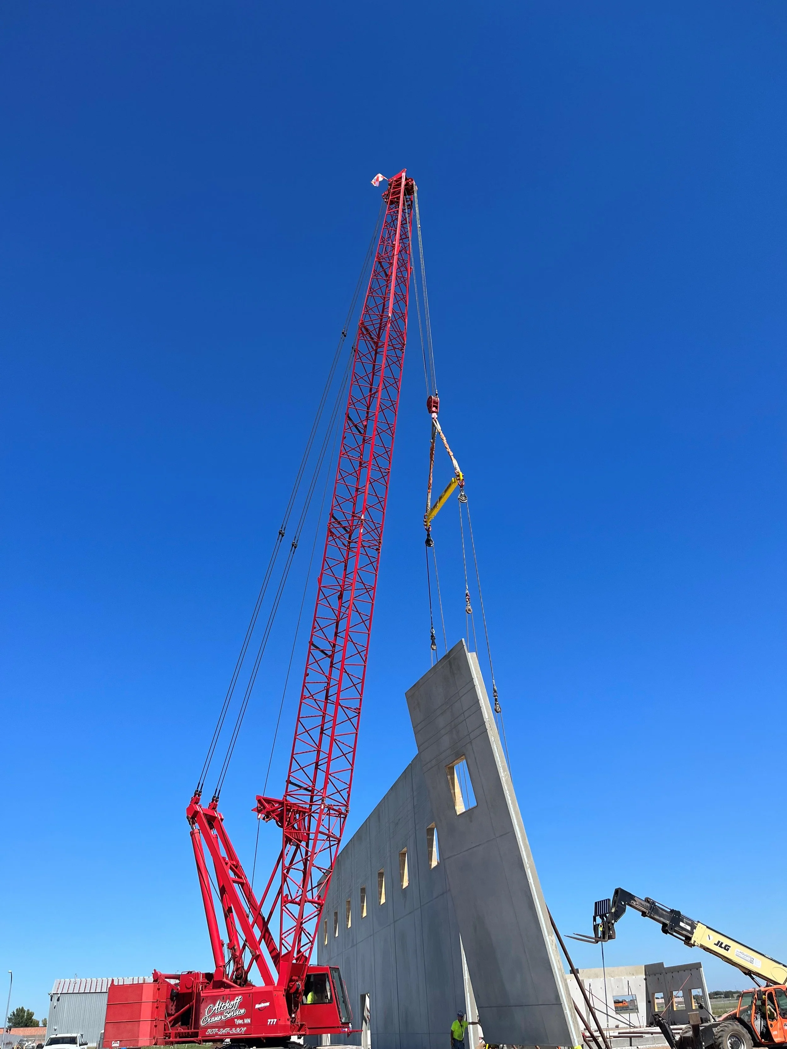 Construction site with a red crane lifting a large concrete wall section into place against a clear blue sky.