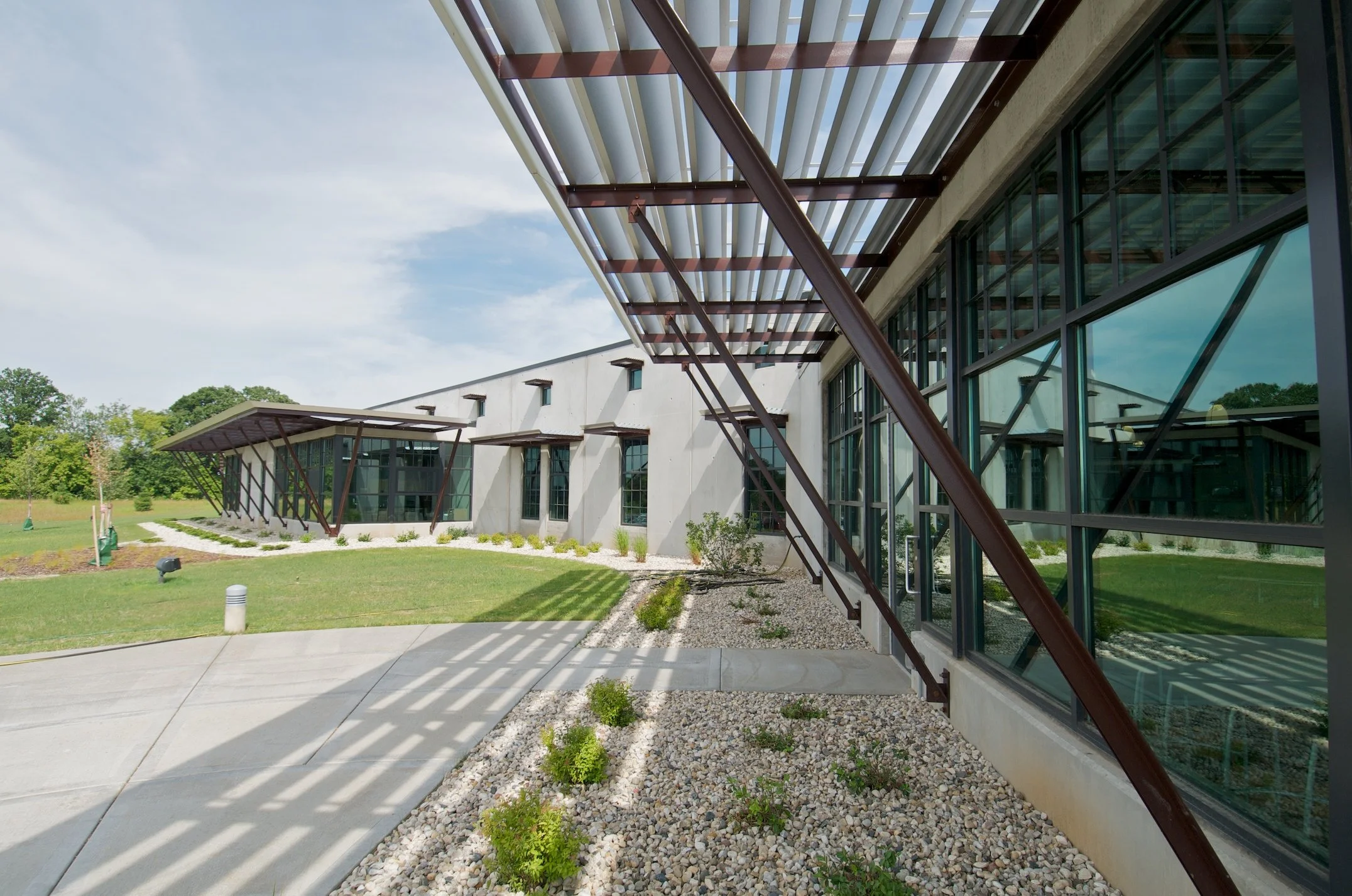 Exterior view of a modern building with large glass windows and metal sunshades, surrounded by a landscaped area with small plants and pathways.