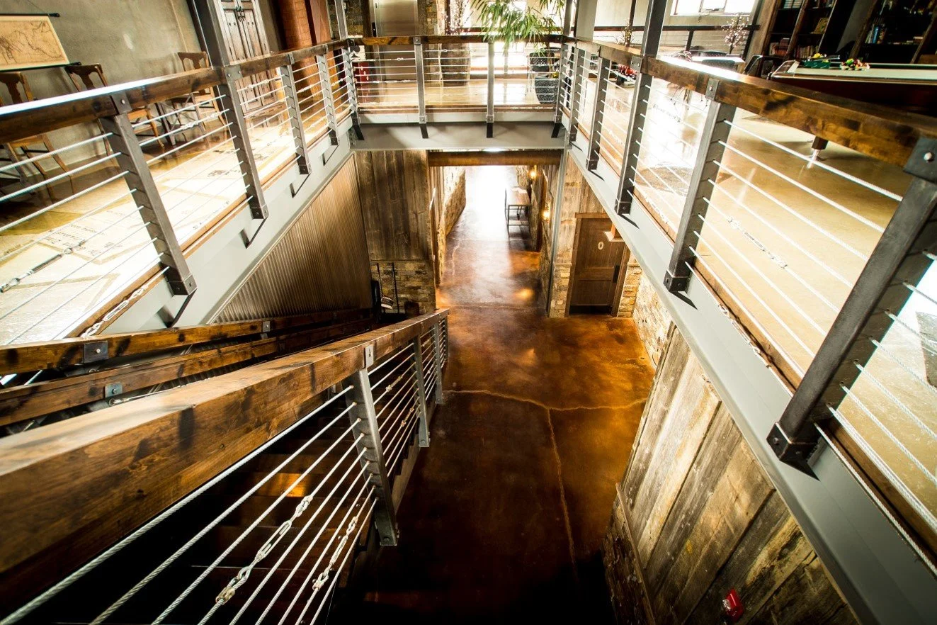 A view of a two-story interior with wooden and metal railing, rustic wood and brick walls, and a polished concrete floor, seen from the second floor looking down.