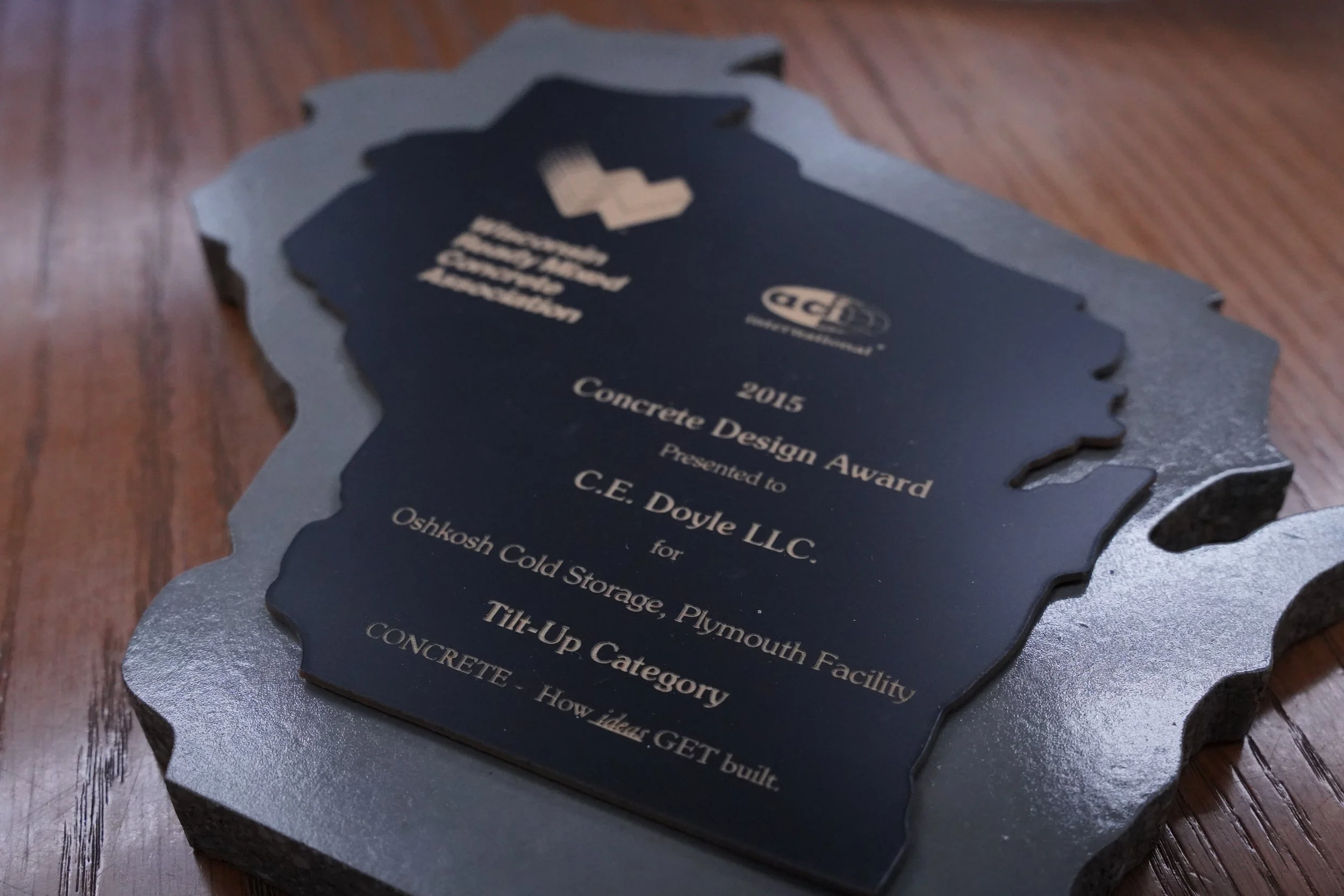 Close-up of a blue and silver award plaque in the shape of a state outline, placed on a wooden surface. The plaque has text that reads '2015 Concrete Design Award Presented to C.E. Doyle LLC for Oshkosh Cold Storage, Plymouth, Tilt-Up Category, How Idaho Gets Built.'