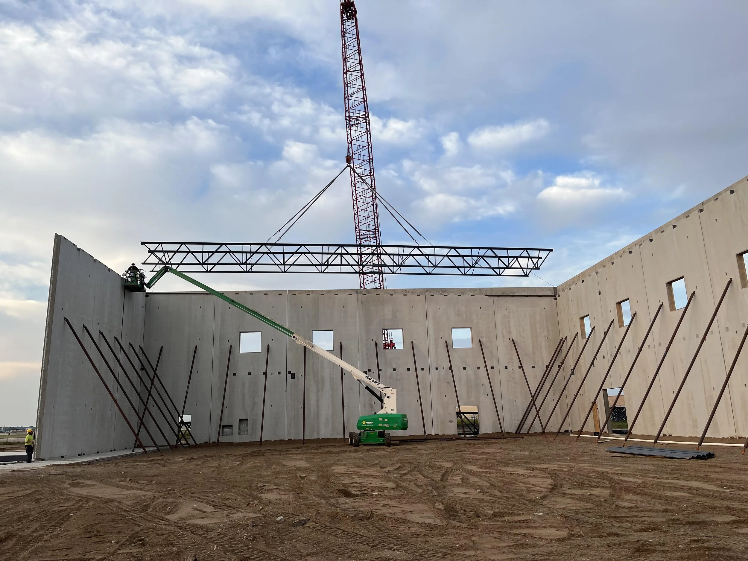 Construction site with a partially built structure, wall panels, and a suspended crane in the background under a cloudy sky.