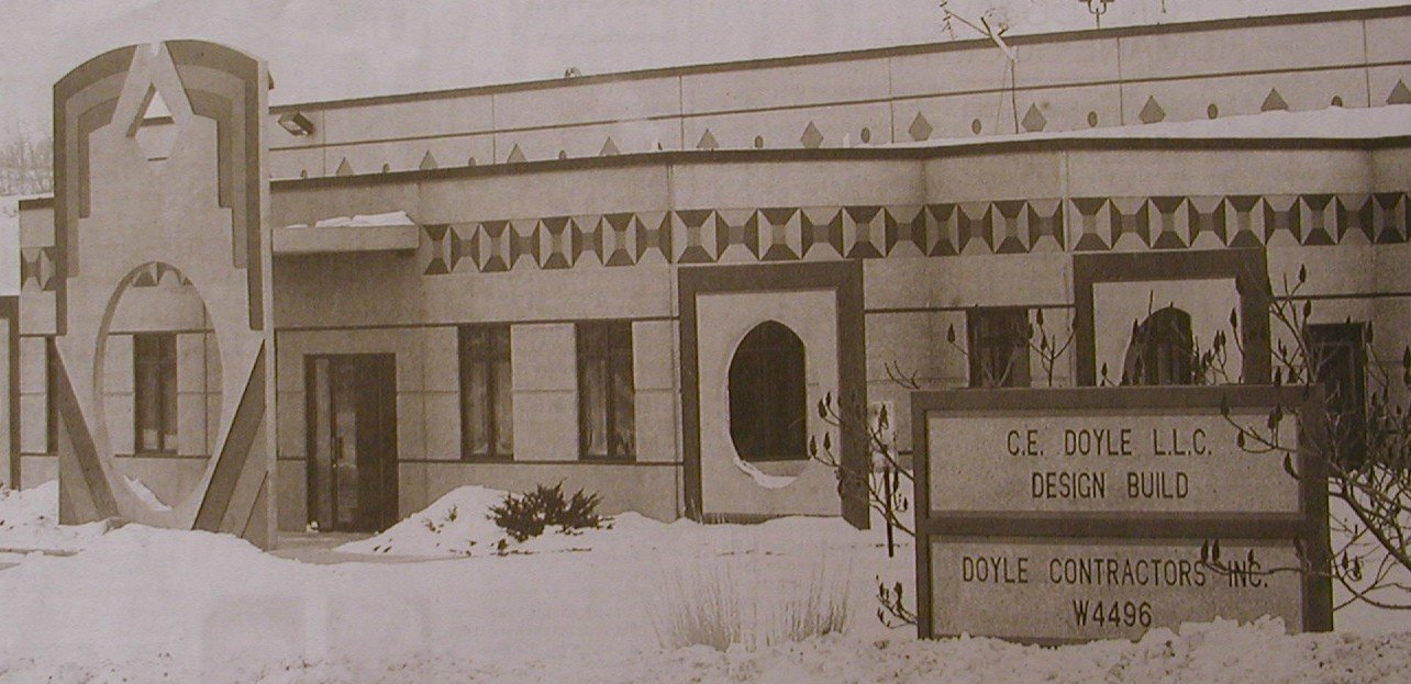 Black and white photo of a building with decorative geometric patterns, a sign stating C.E. Doyle L.L.C. Design Build, Doyle Contractors Inc., W4496, and some snow-covered landscape.