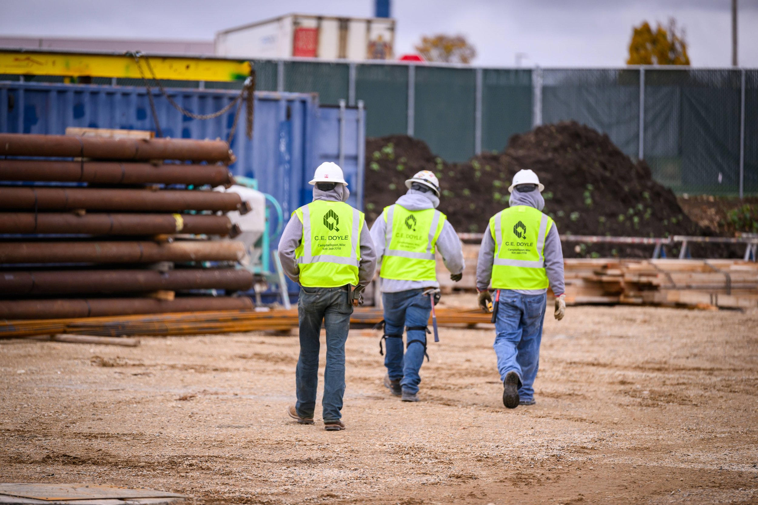 Three construction workers wearing bright yellow safety vests and white helmets walk on a dirt construction site, with metal pipes and a blue container in the background.