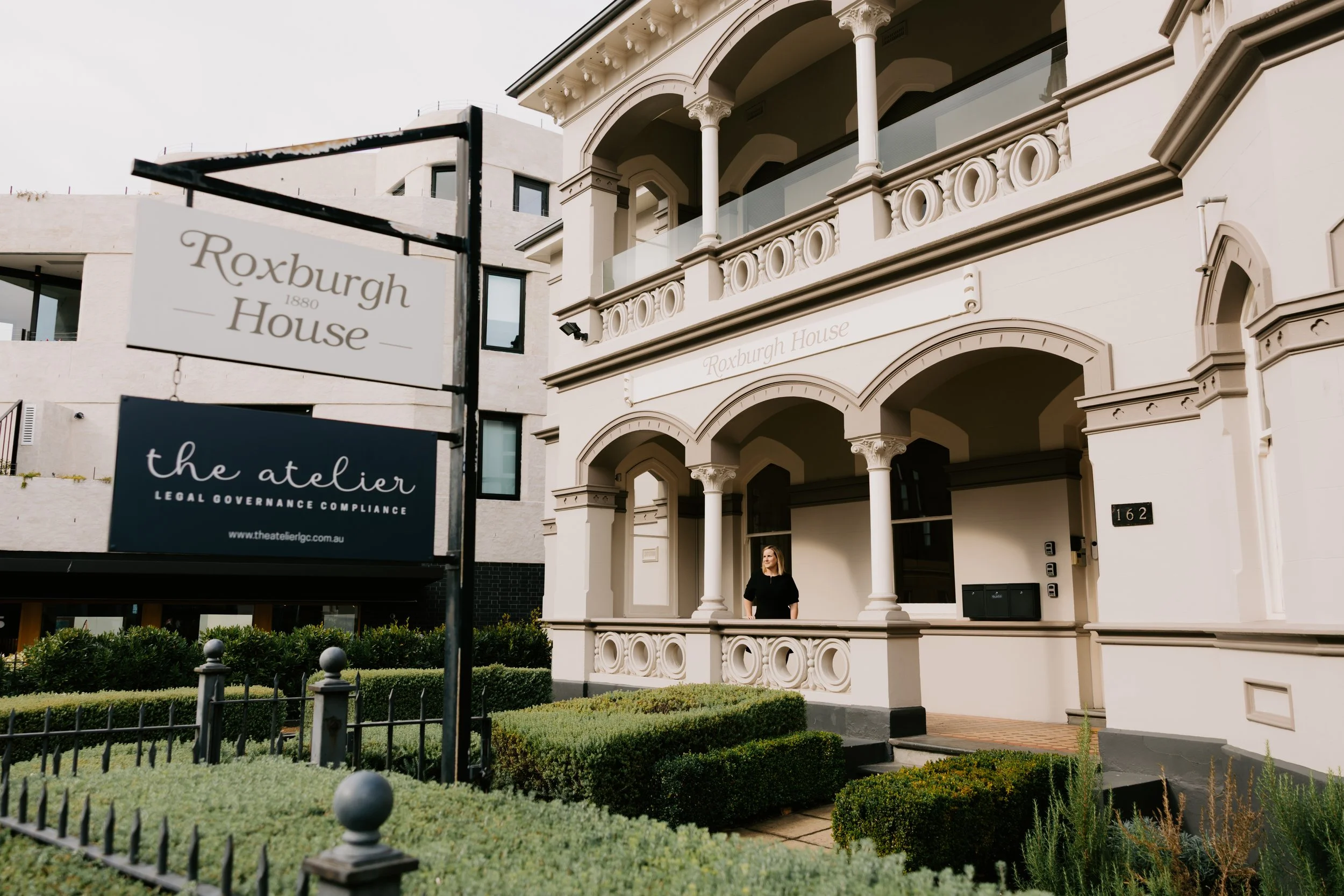 A woman standing on the porch of a white, ornate Victorian-style house, with signs in the foreground reading ‘Roxburgh House 1880’ and ‘the atelier Legal Governance Compliance’. The house has decorative columns and balustrades, and there is well-maintained greenery in the front yard.