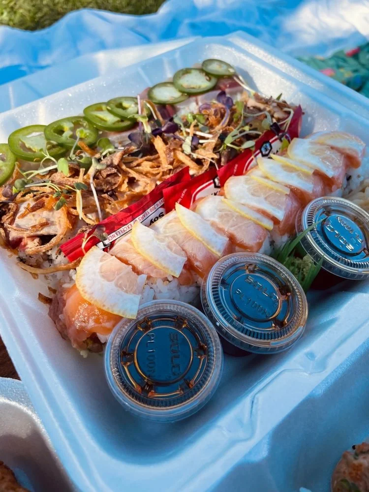 A styrofoam container holding sushi rolls topped with lemon slices, slices of salmon, and small containers of soy sauce, with a side of wasabi and ginger, outdoors on a blue surface.