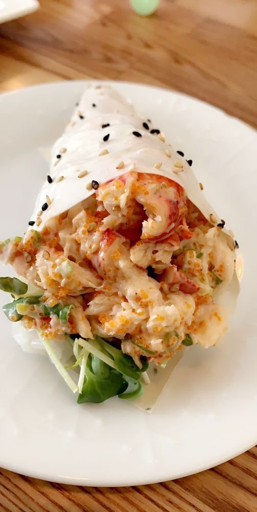 A sushi roll topped with spicy seafood salad, garnished with white and black sesame seeds, served on a white plate with some greens underneath.