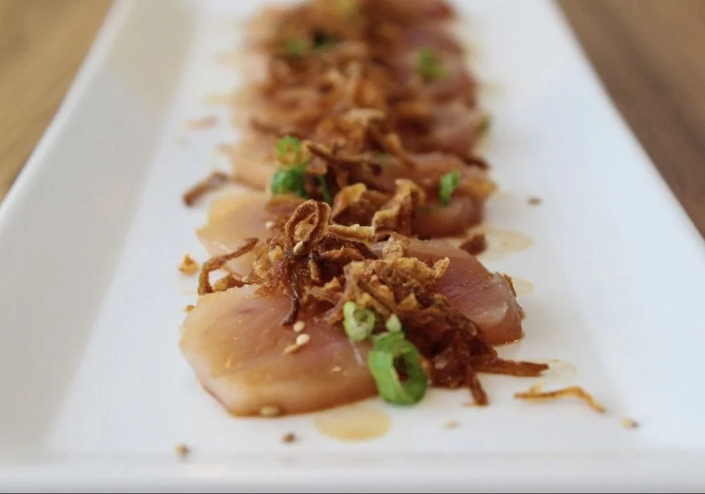 Close-up of sliced raw fish, topped with crispy fried shallots and chopped green onions, served on a white rectangular plate.