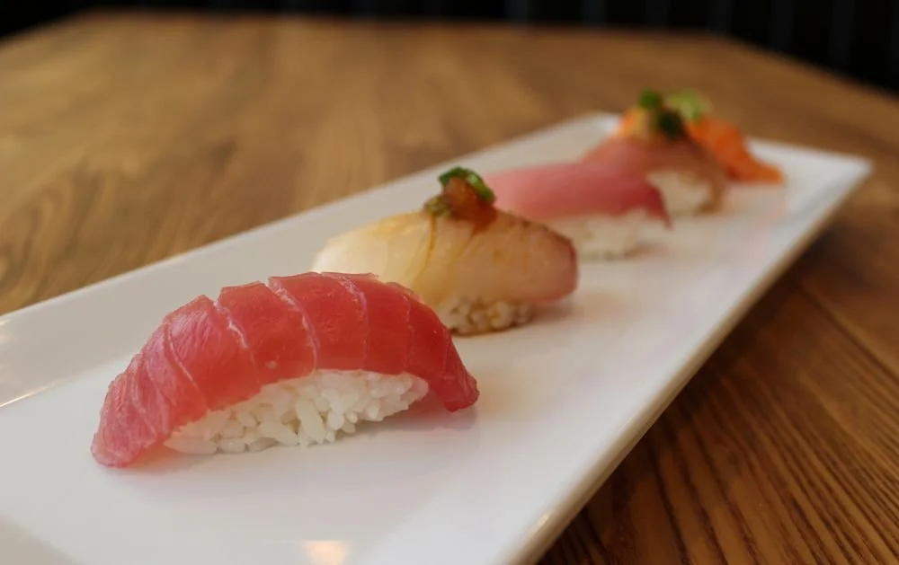 Four pieces of sushi with various fish on white rice, served on a rectangular white plate on a wooden table.