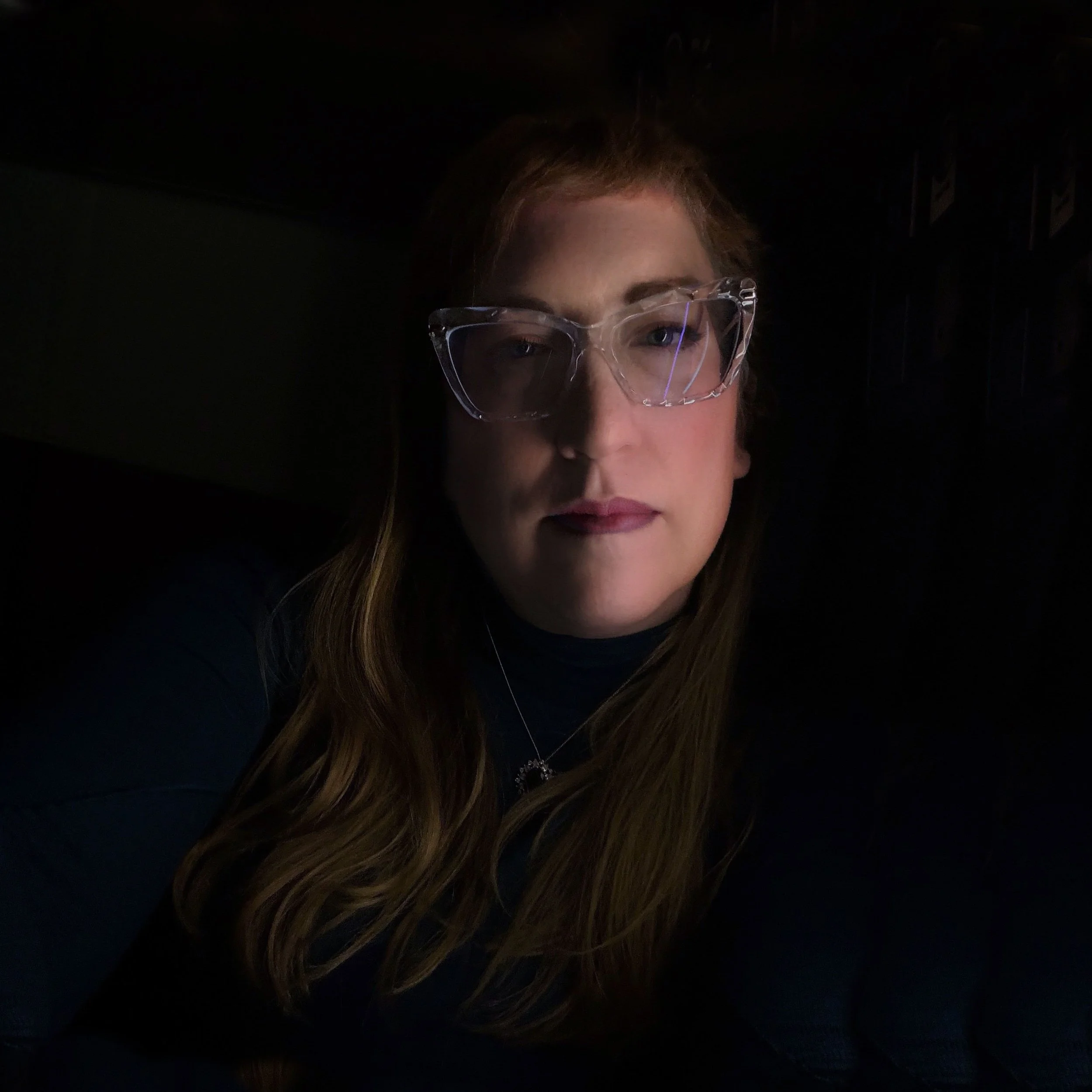 A woman with long brown hair wearing clear glasses and a dark top, in a dimly lit environment.