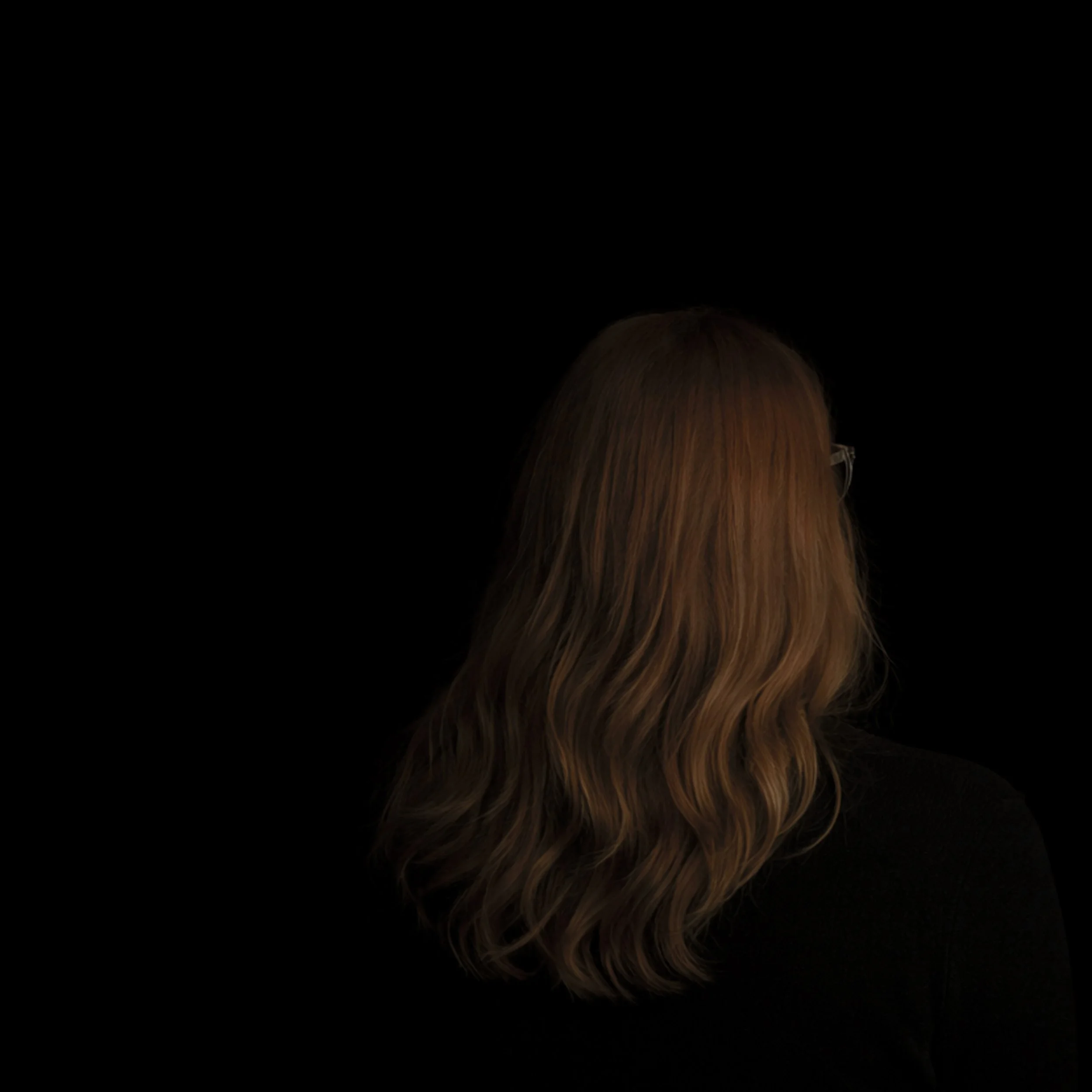 Back view of a woman with long wavy auburn hair, wearing glasses and a black top, against a plain black background.