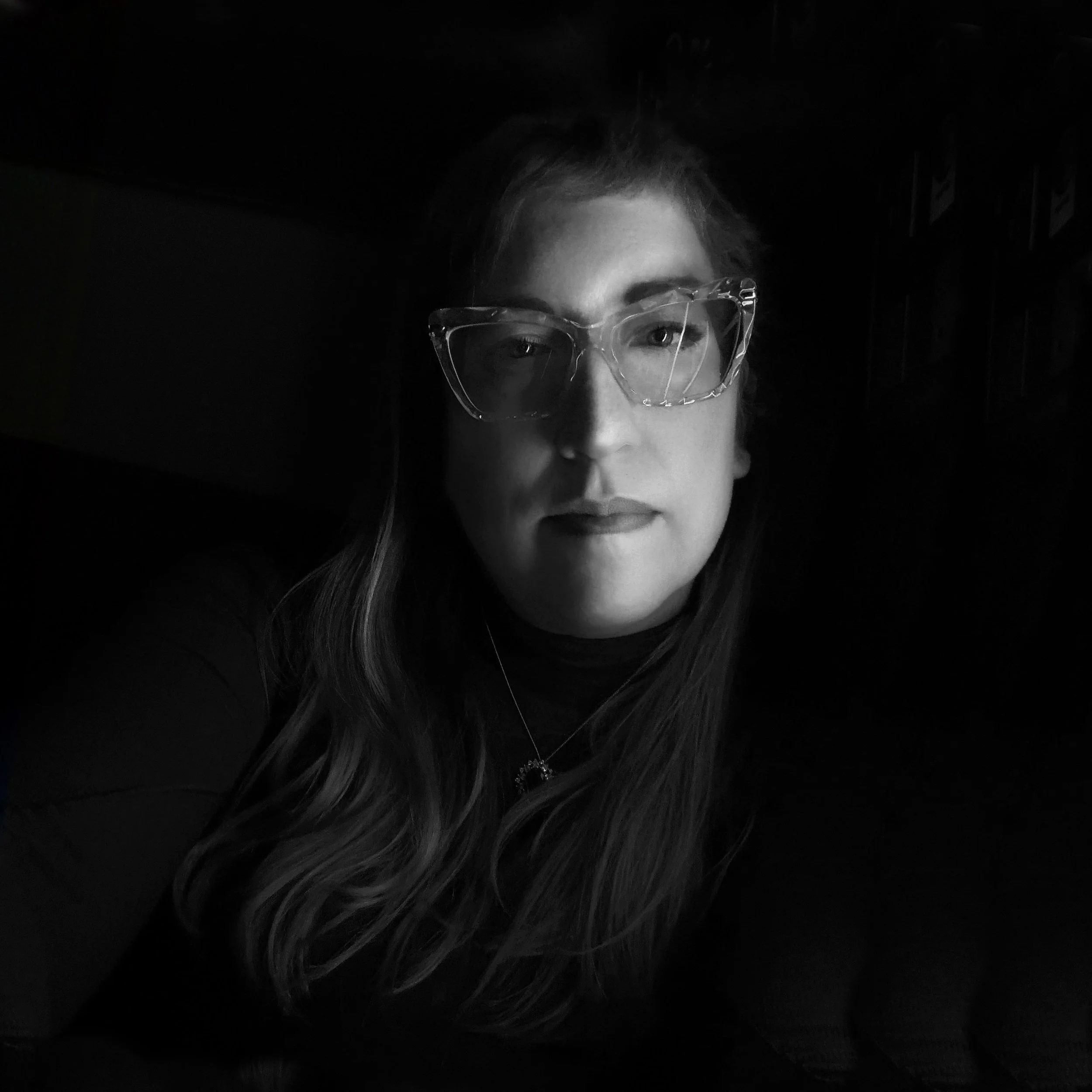 A woman with long hair wearing glasses and a necklace, looking into the camera in a dimly lit setting.