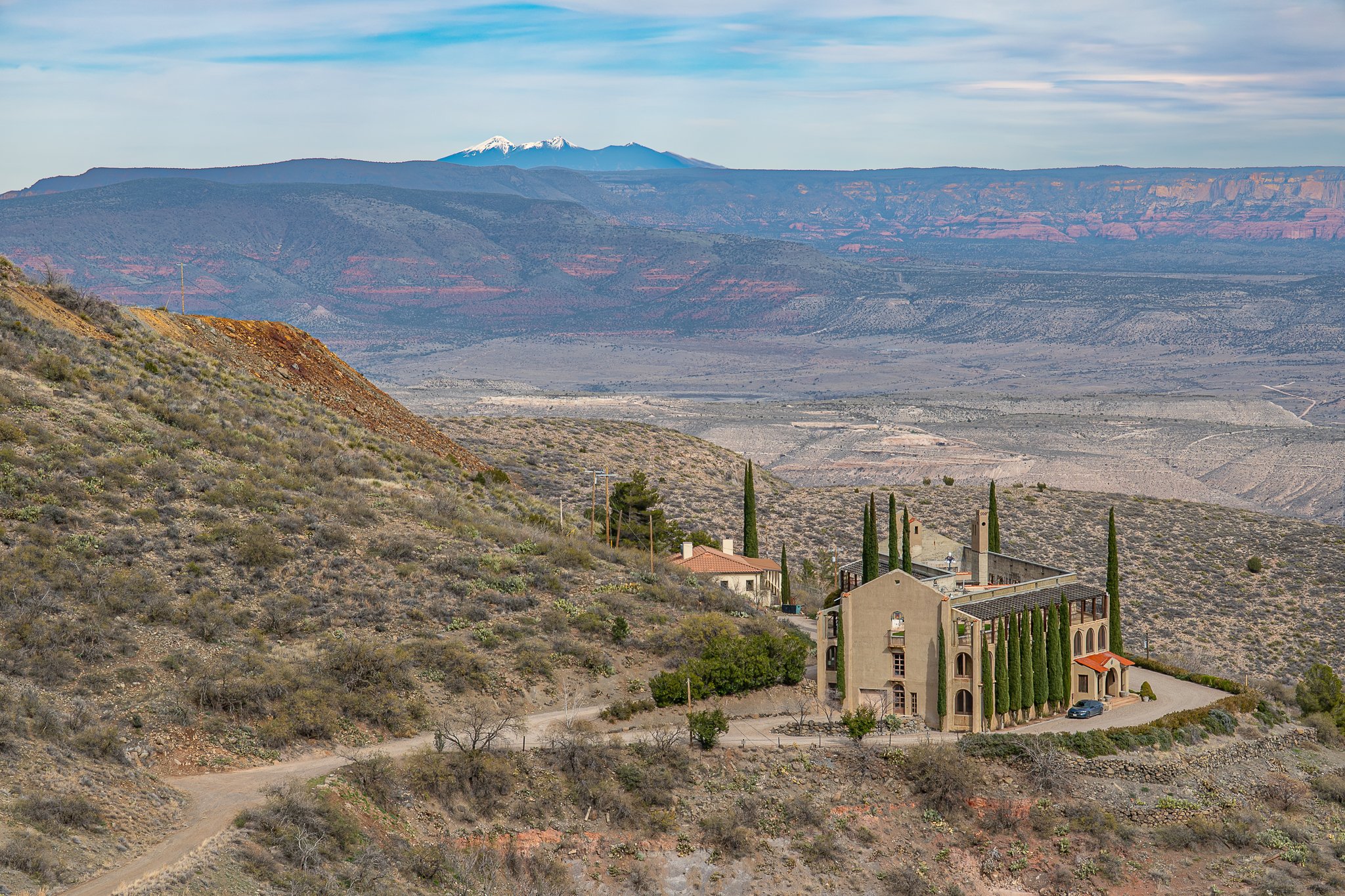 A house in the hills with tall cypress trees, overlooking a desert valley, with mountains in the background and snow-capped peaks in the distance.
