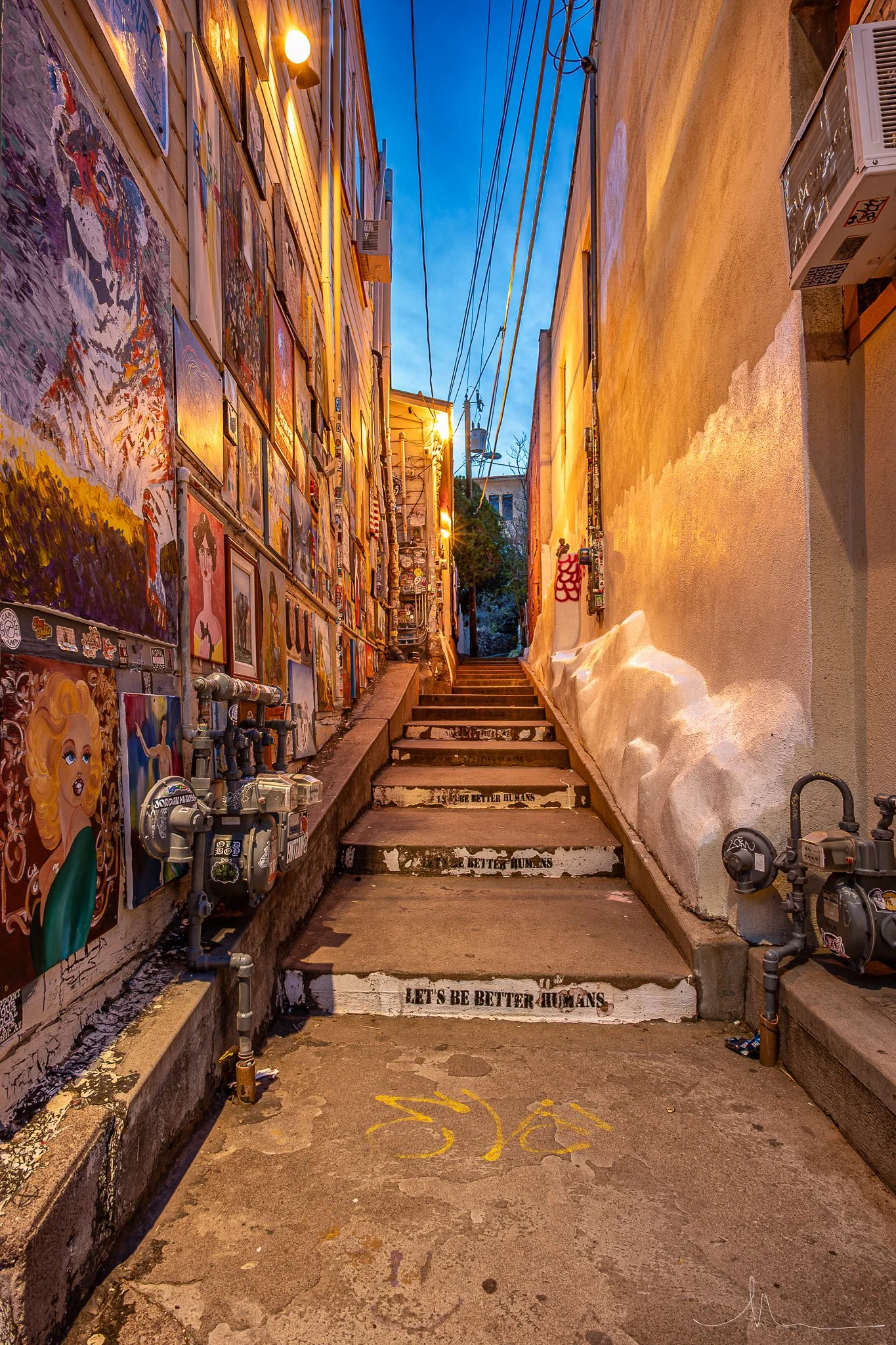 Street alleyway with colorful art on the left wall, street lights, stairs leading upward, electrical meters and wires overhead, and a painted bicycle symbol on the pavement in the foreground.