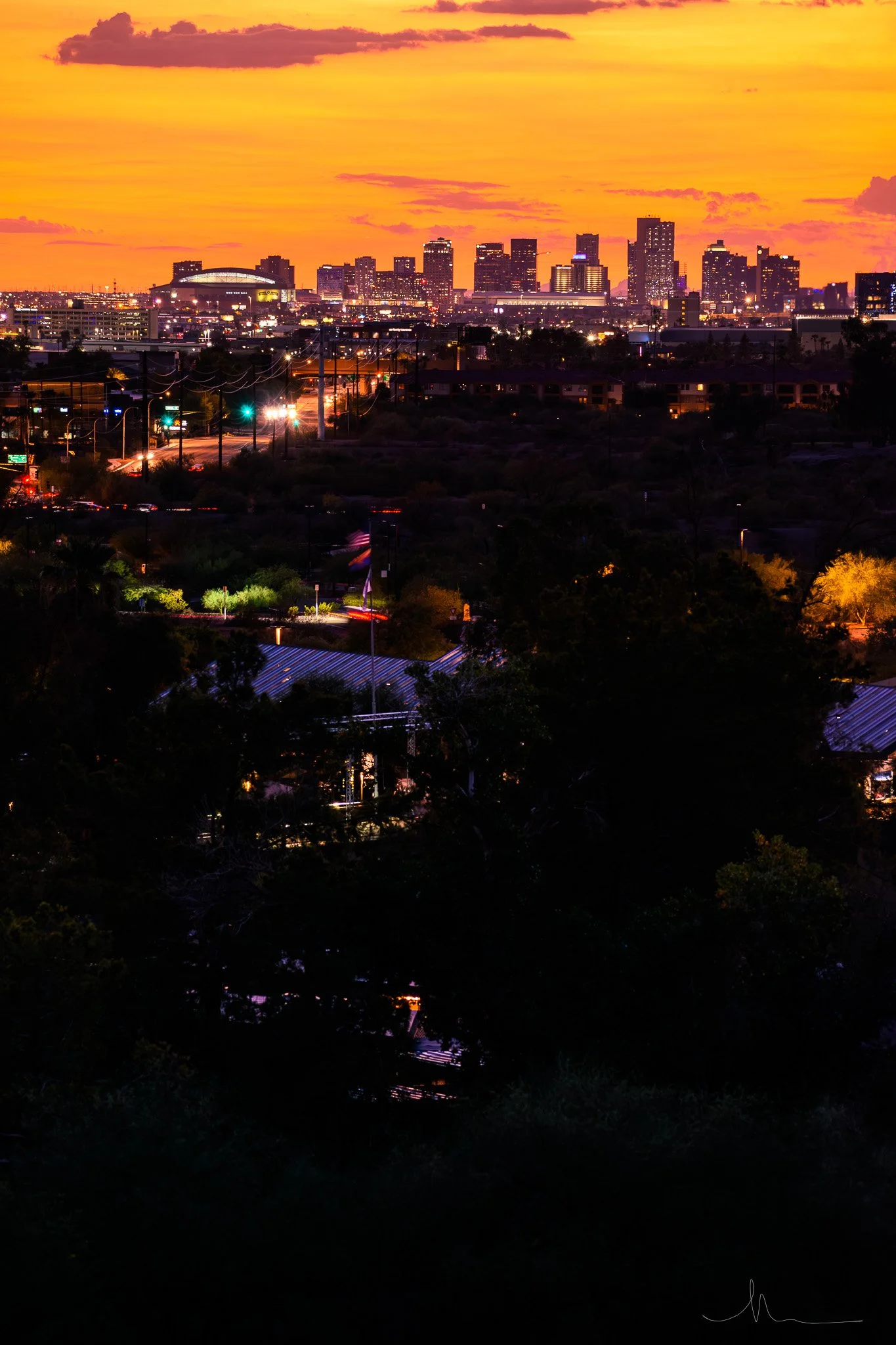 Nighttime cityscape of a downtown area with illuminated buildings, streetlights, and a vibrant sunset sky in the background.