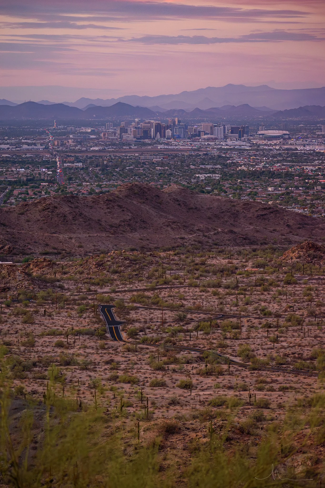 A desert landscape in the foreground with sparse vegetation and a winding paved road. The middle ground features rocky hills and mountains, with a city skyline and mountains in the distance under a purple and pink sky.