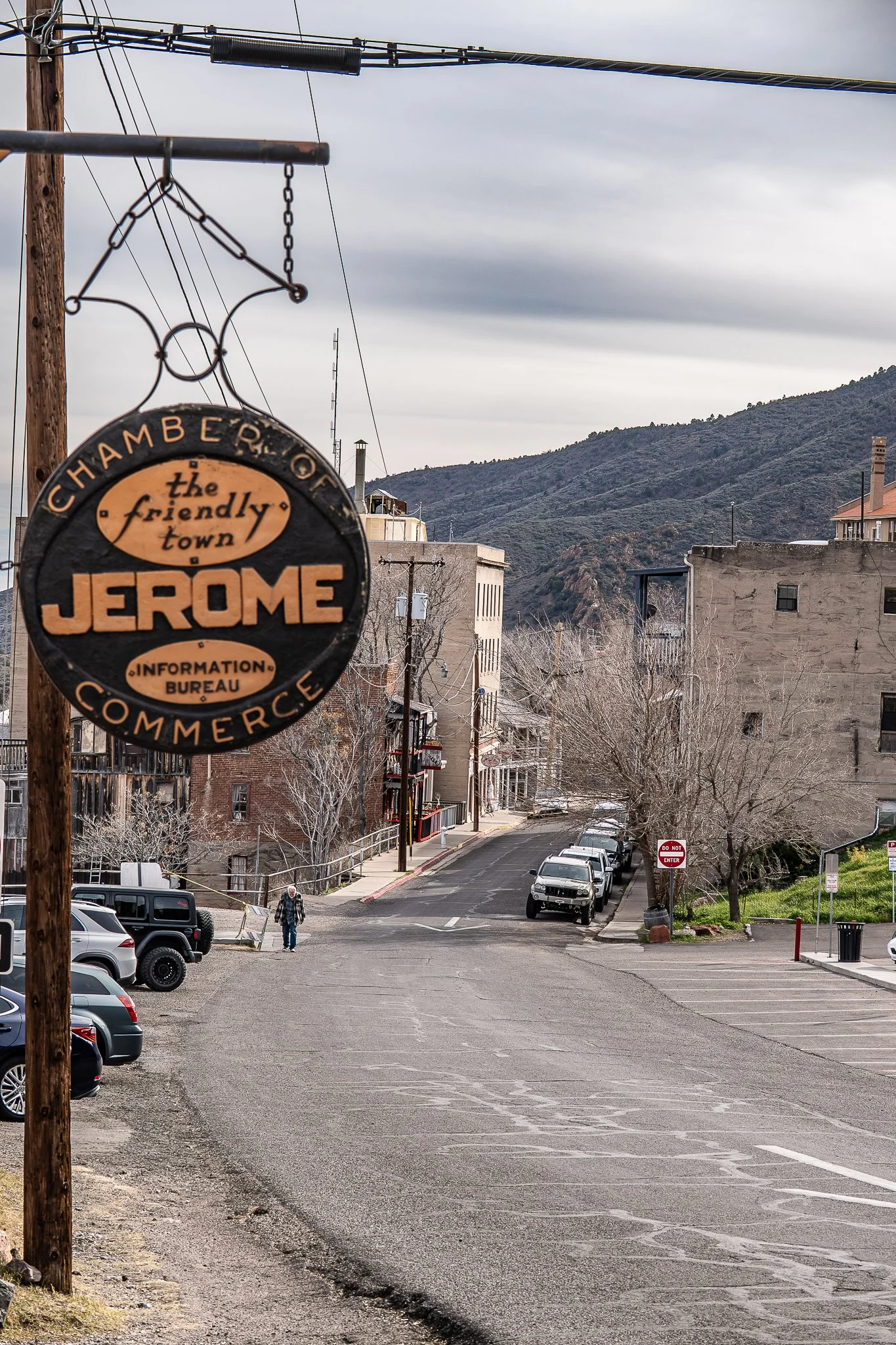 A street scene in Jerome, with a hanging sign for the Chamber of Commerce in the foreground. The street curves uphill, lined with parked cars and, some leafless trees, and multi-story buildings. Mountains are visible in the background under an overca