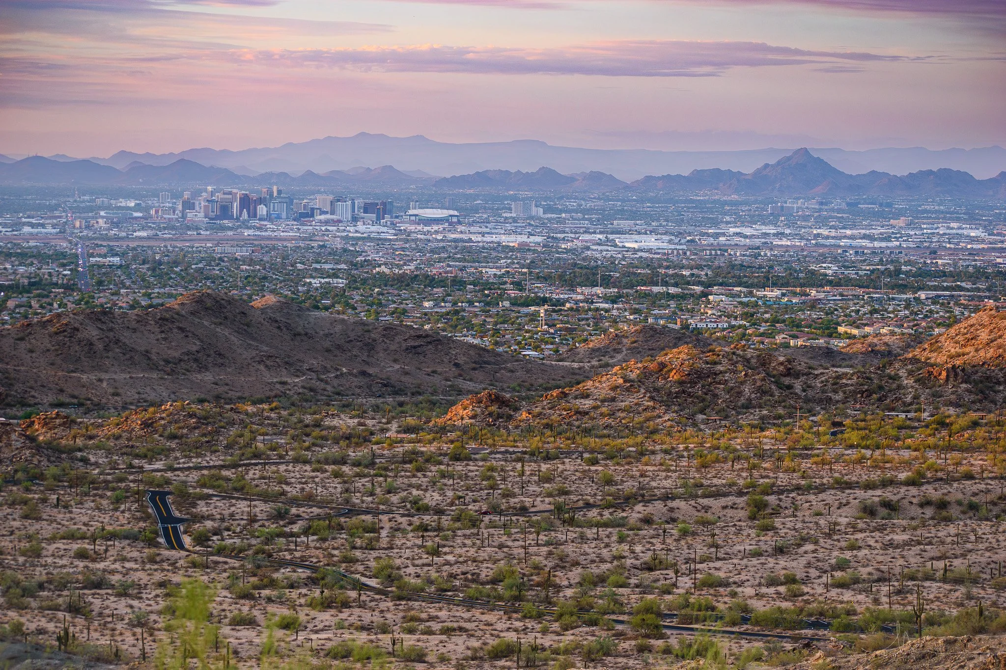 Scenic view of a desert landscape with mountains, sparse vegetation, and winding roads in the foreground, and a city skyline with high-rise buildings in the distance under a pink and purple sunset sky.
