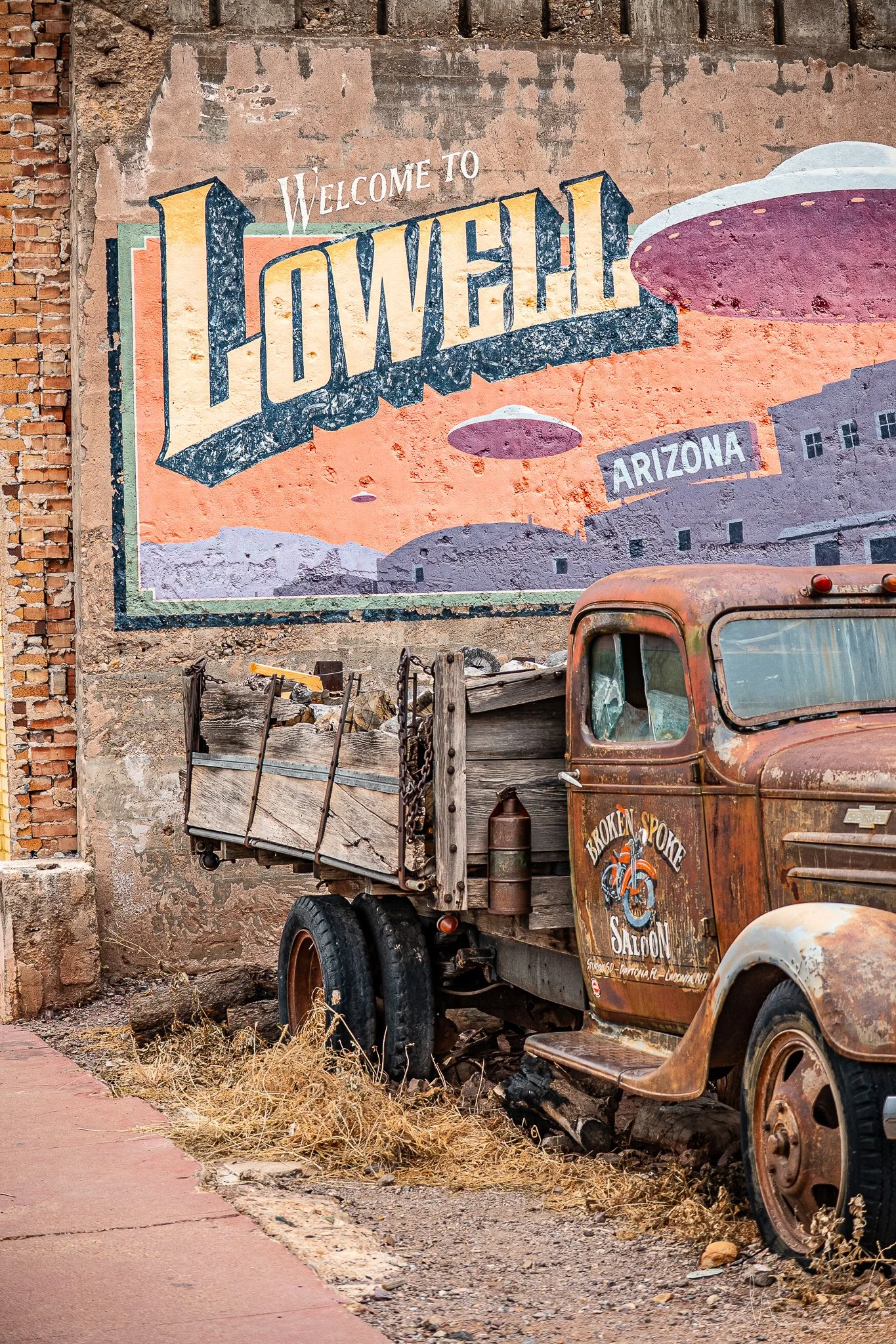 A vintage rusted truck parked below a colorful mural that reads 'Welcome to Lowell, Arizona' with a desert landscape and flying saucers painted on the wall.