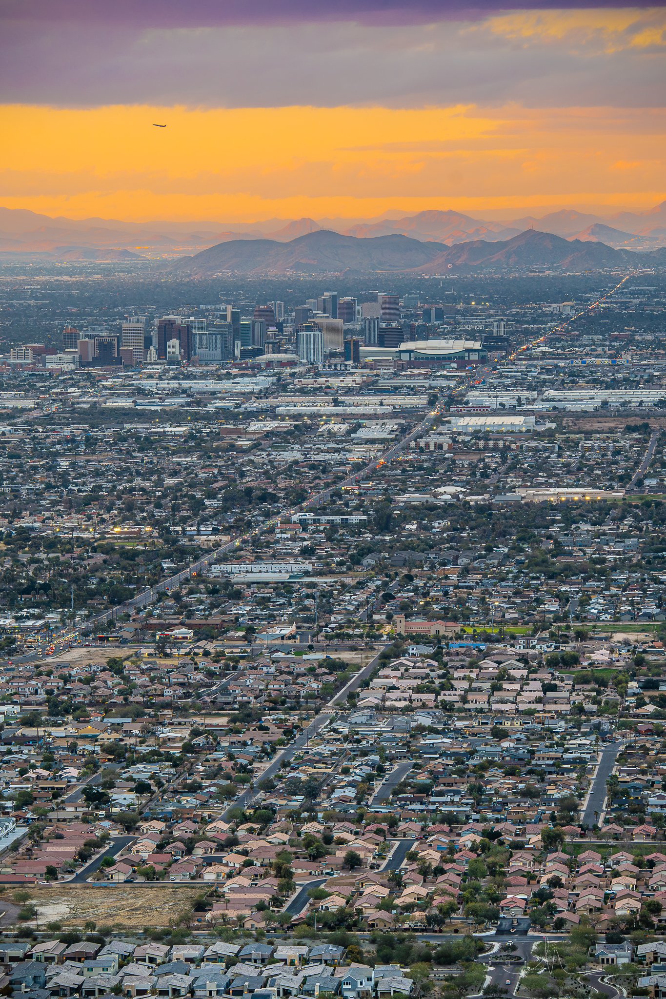 An aerial view of a cityscape at sunset, showing downtown skyscrapers, residential neighborhoods, and mountain ranges in the background.