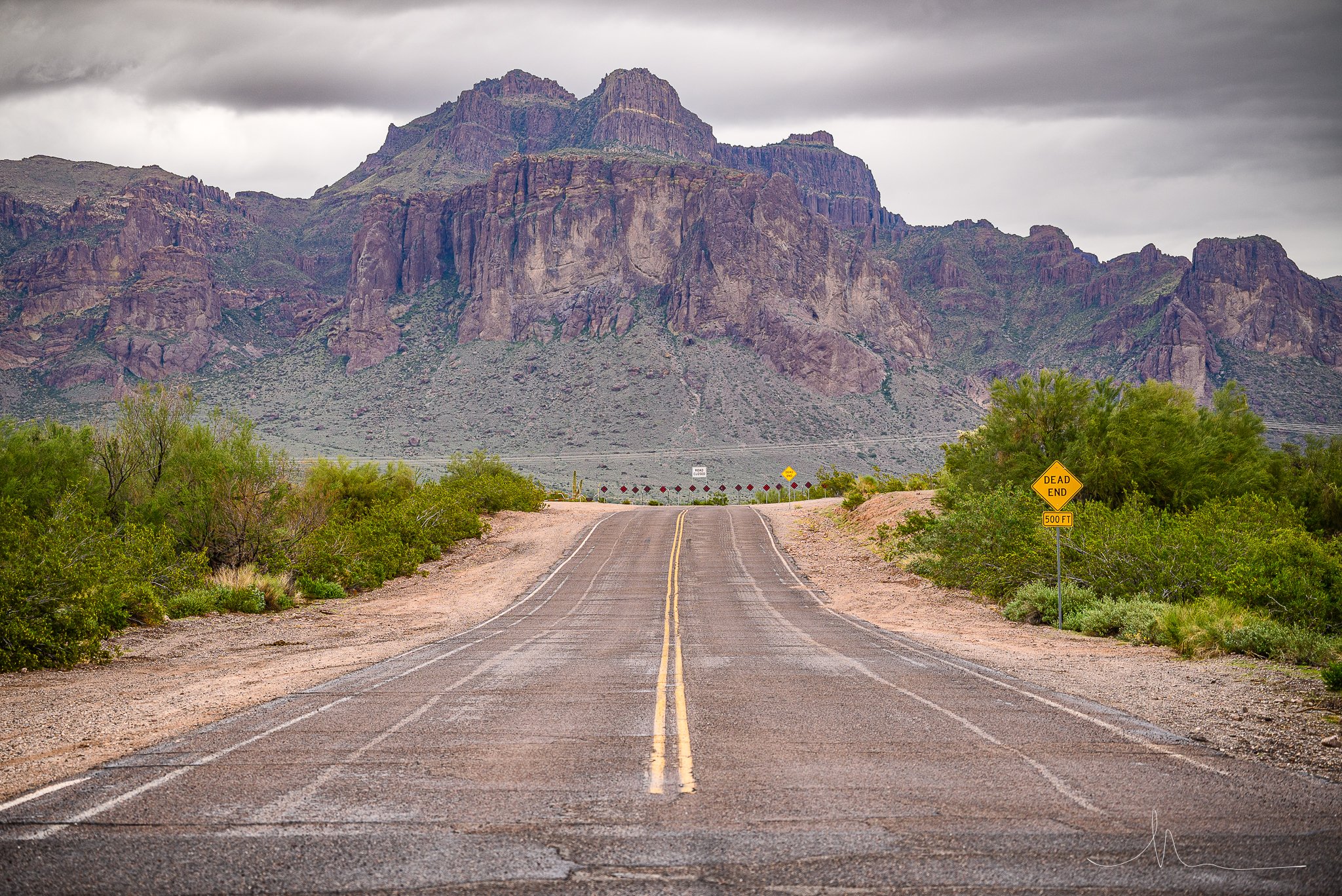 A deserted desert road leading towards mountains under cloudy sky, with a yellow 'Dead End' sign on the right side.