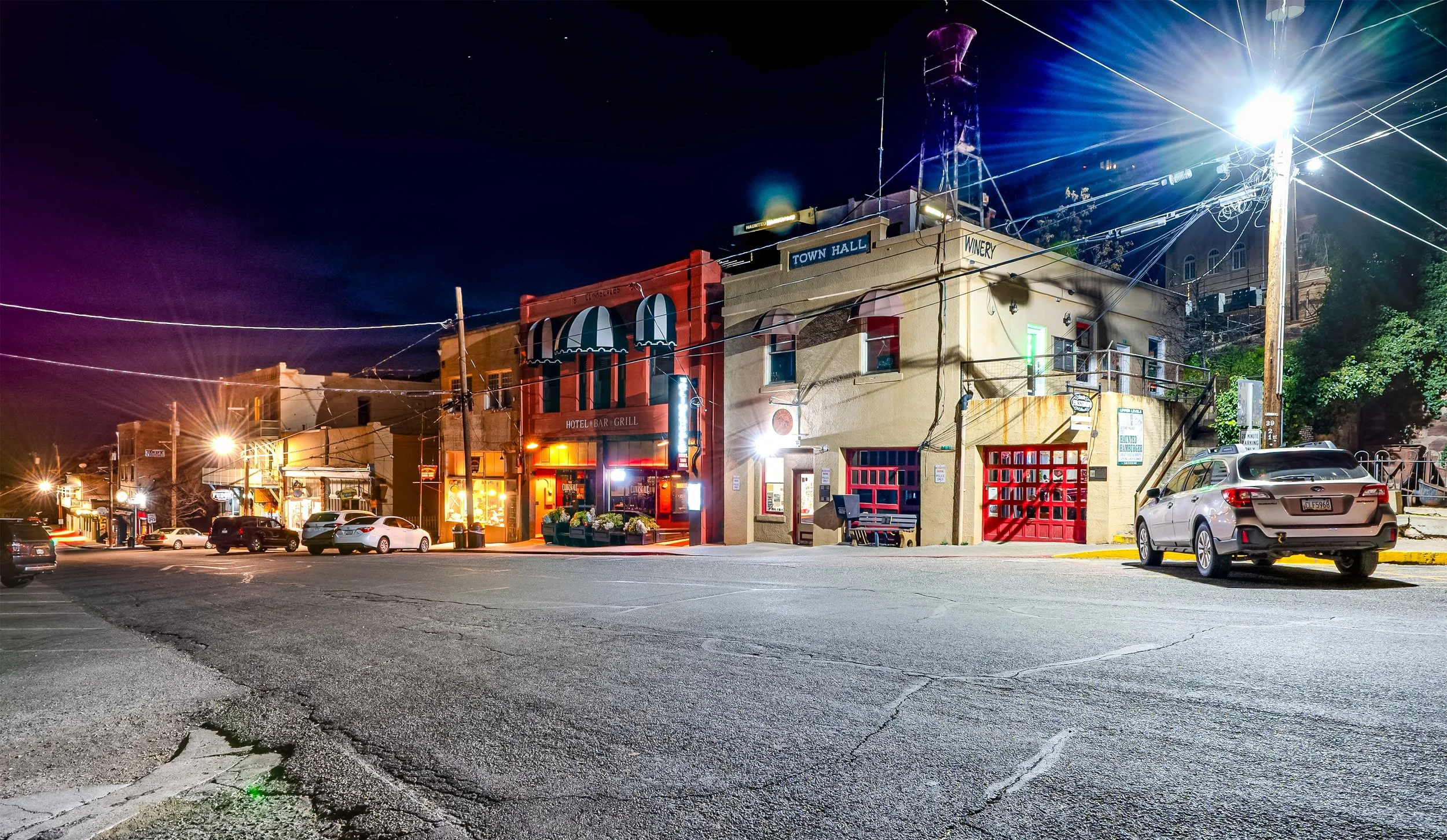 Nighttime view of a small town street with lit storefronts and cars parked along the road. Buildings include a hotel, bar, grill, town hall, and winery, with power lines overhead and a bright streetlight.
