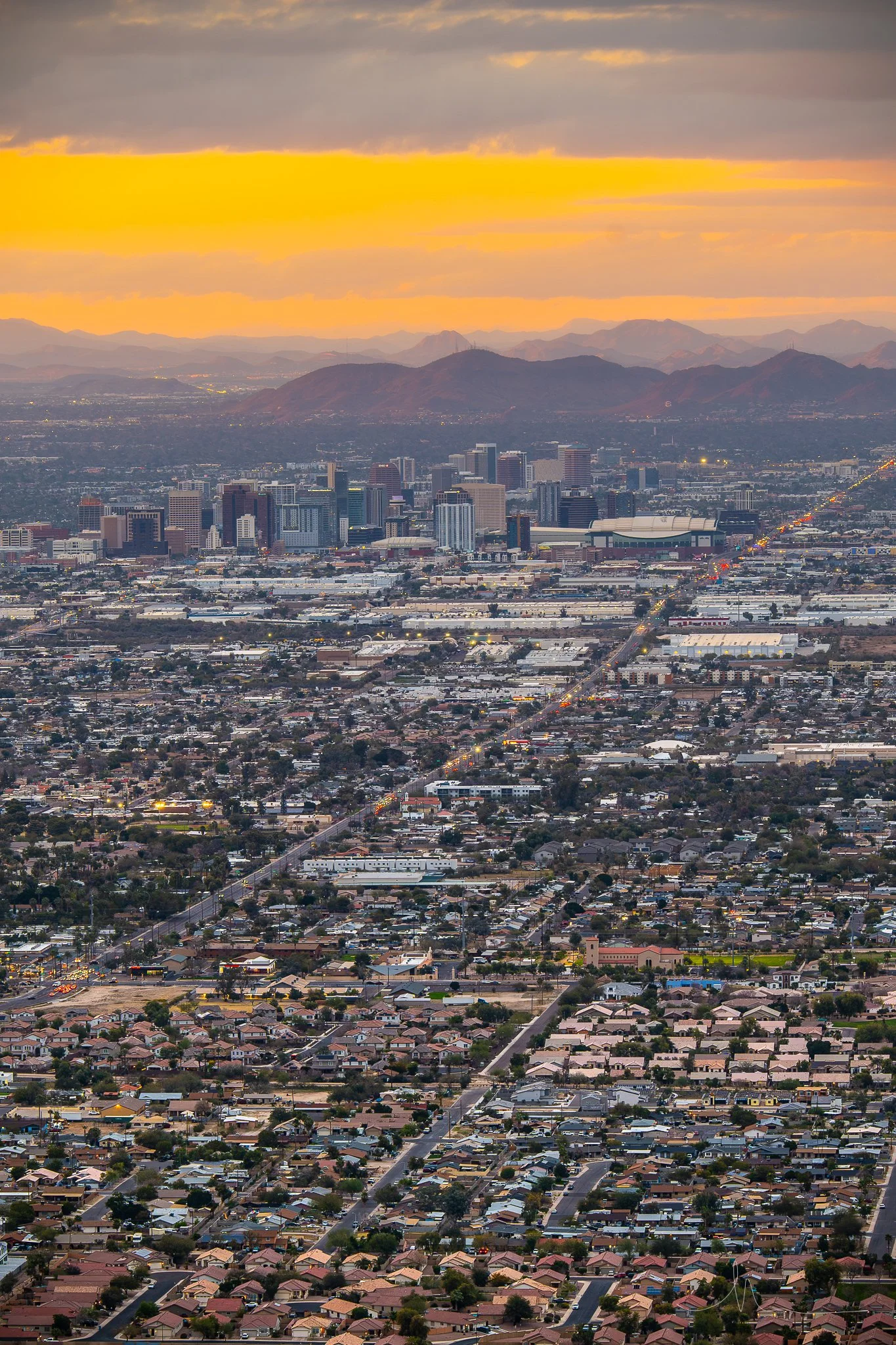 Aerial view of a city skyline during sunset, with tall buildings, suburban neighborhoods, and distant mountains.