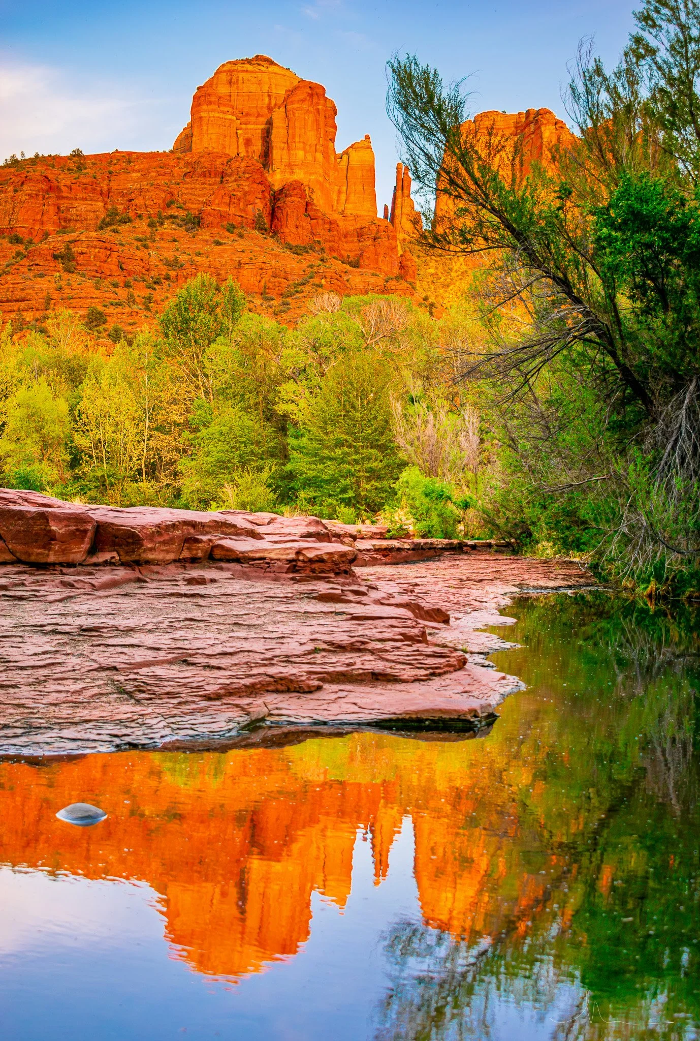 Sunset over red rock formations and a calm river, with green trees in the foreground.