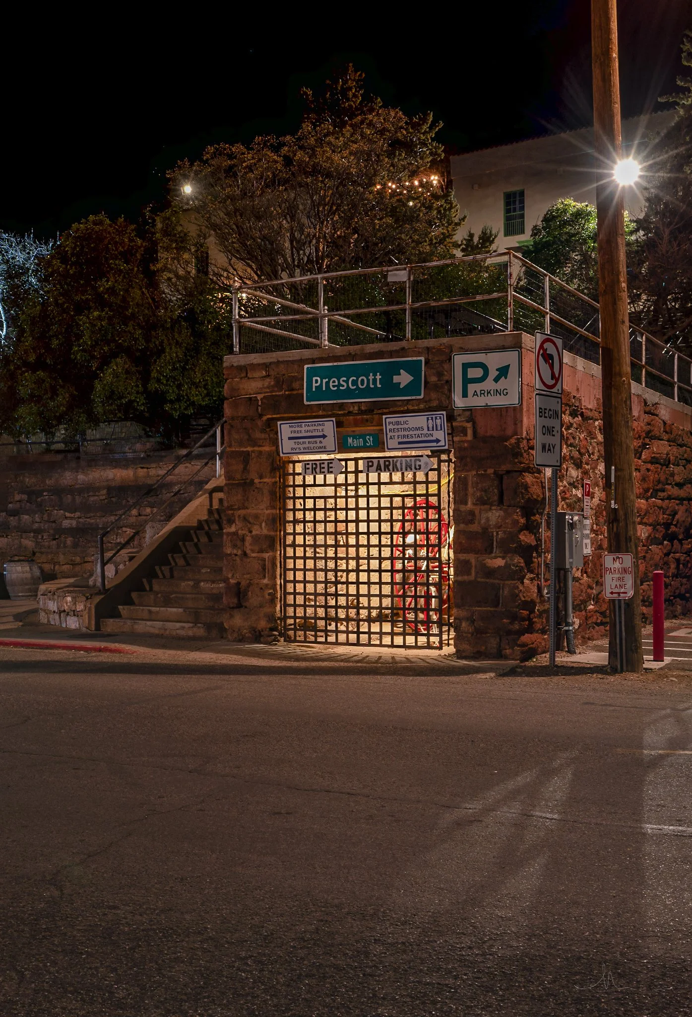 Nighttime street scene showing parking signs, stairs leading up to a platform, and a gated area with a light inside, located near a brick wall and trees.